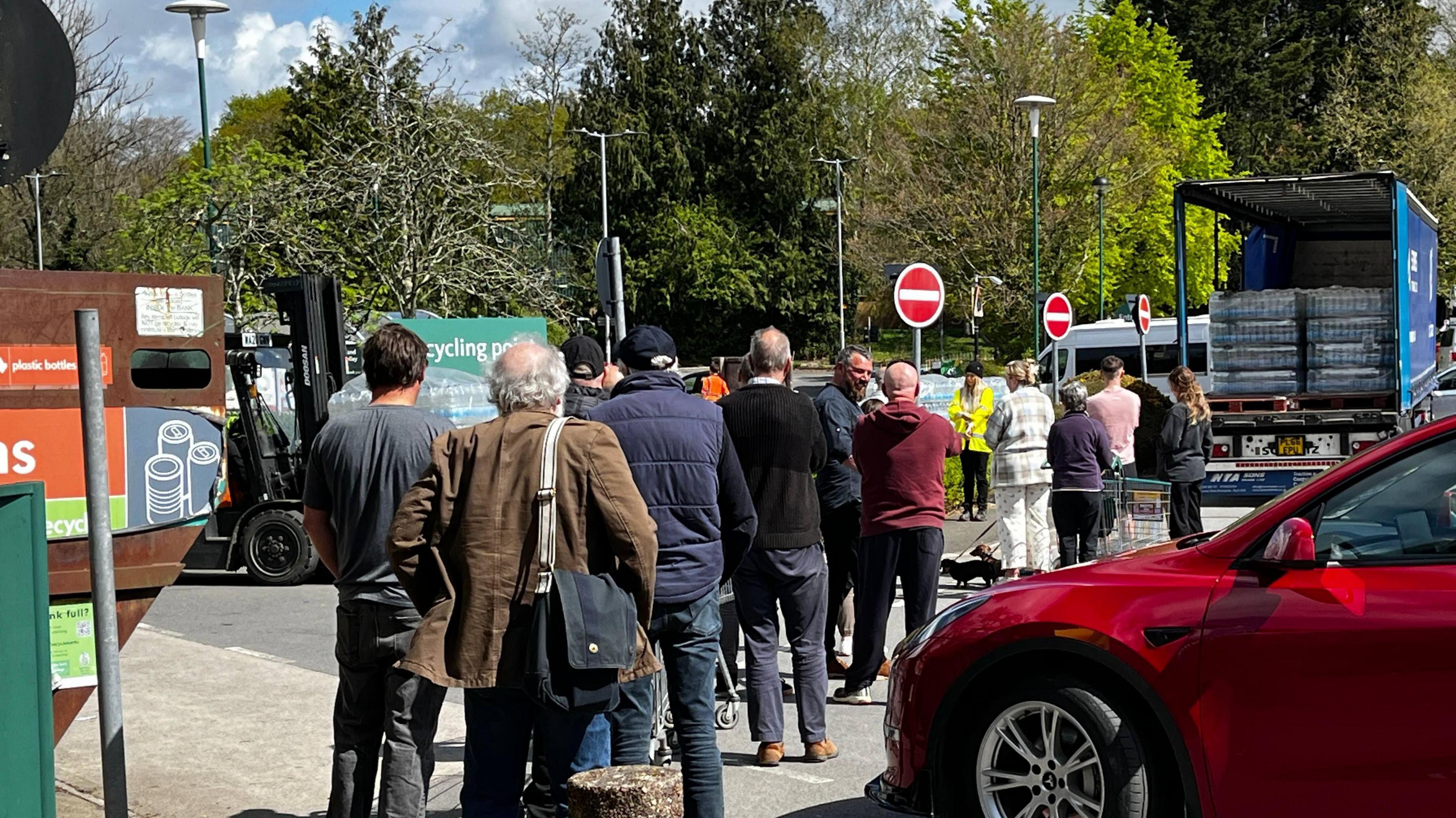 People queuing at a water station in Tavistock. There is a lorry filled with crates of bottled water in the distance with a person wearing hi-vis stood next to it. On the right, there is a parked red car.