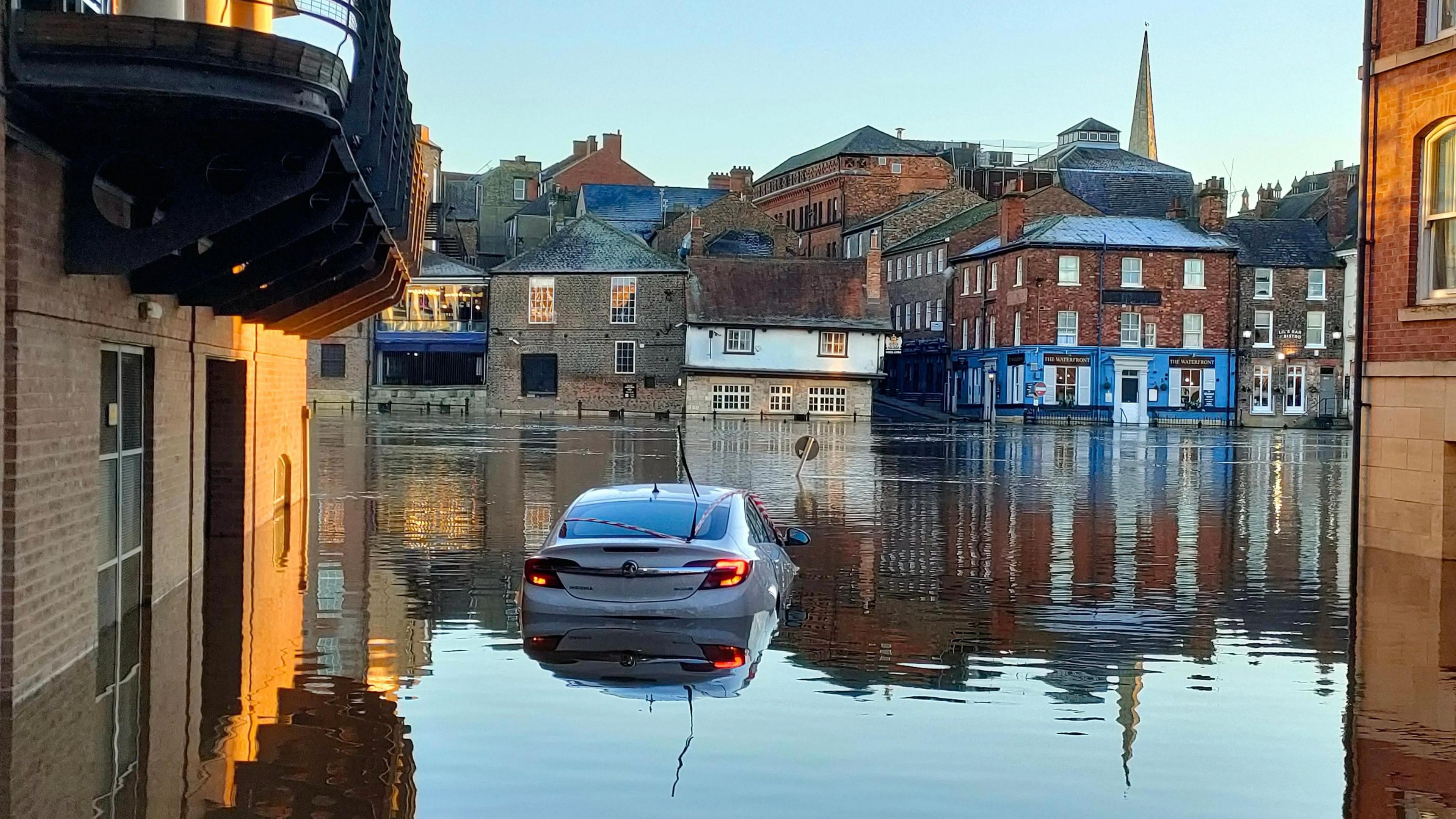 A car is part submerged in flood water from the River Ouse. Buildings can be seen on the opposite bank of the river.