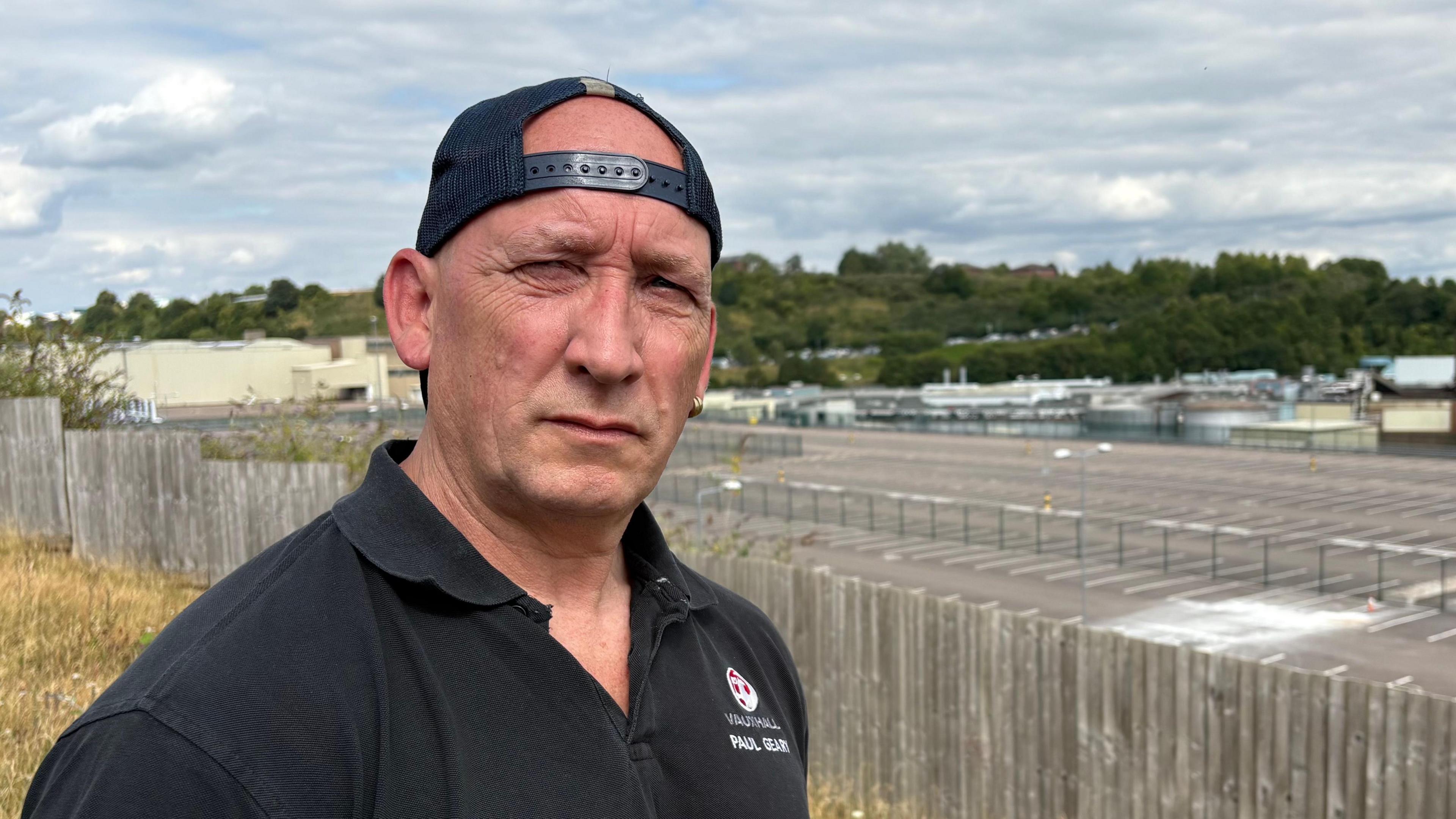 Paul Geary, slightly to the left of frame and looking direct to camera, with his shoulders turned slightly away from us. He is wearing a black cap, which is turned backwards, and a black T-shirt embroidered with the red and white Vauxhall logo and Paul's name, which are both just visible. He is standing outside, in front of an empty car park. It is a bright day but there are lots of clouds in the sky.