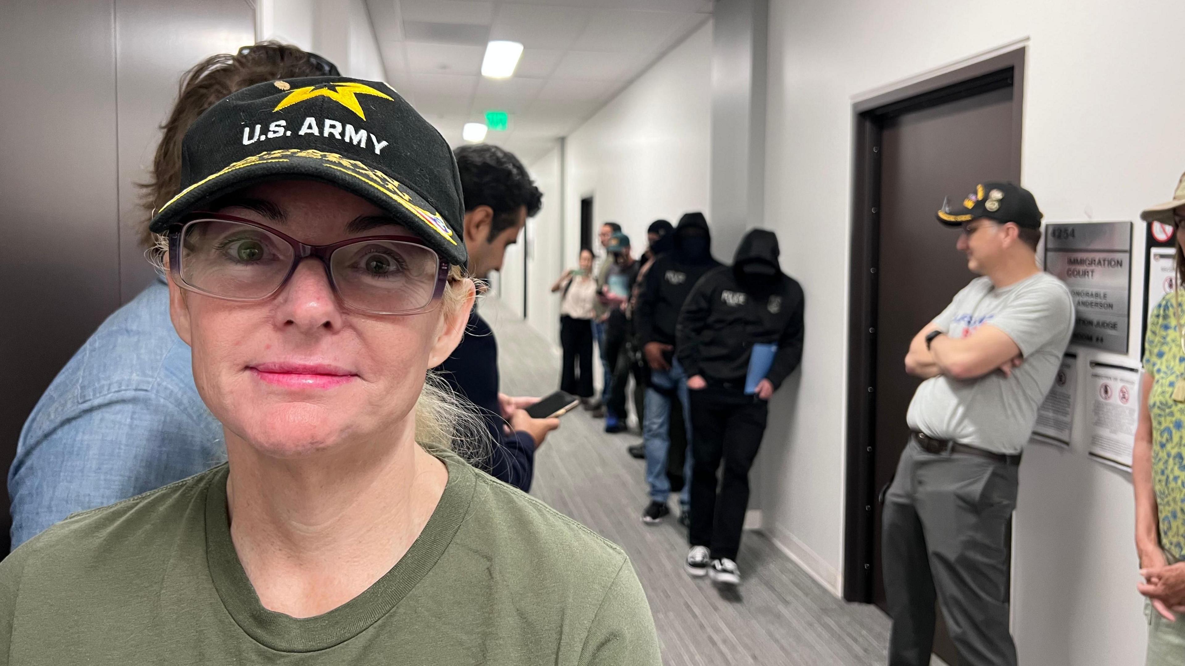 Monique stares at the camera, wearing a black baseball cap that says US Army, and an olive-green t-shirt. In the background is a fluorescent hallway and a man in a gret t-shirt and the same black army cap. Several ICE officers, masked and in black hoodies, are also in the hallway