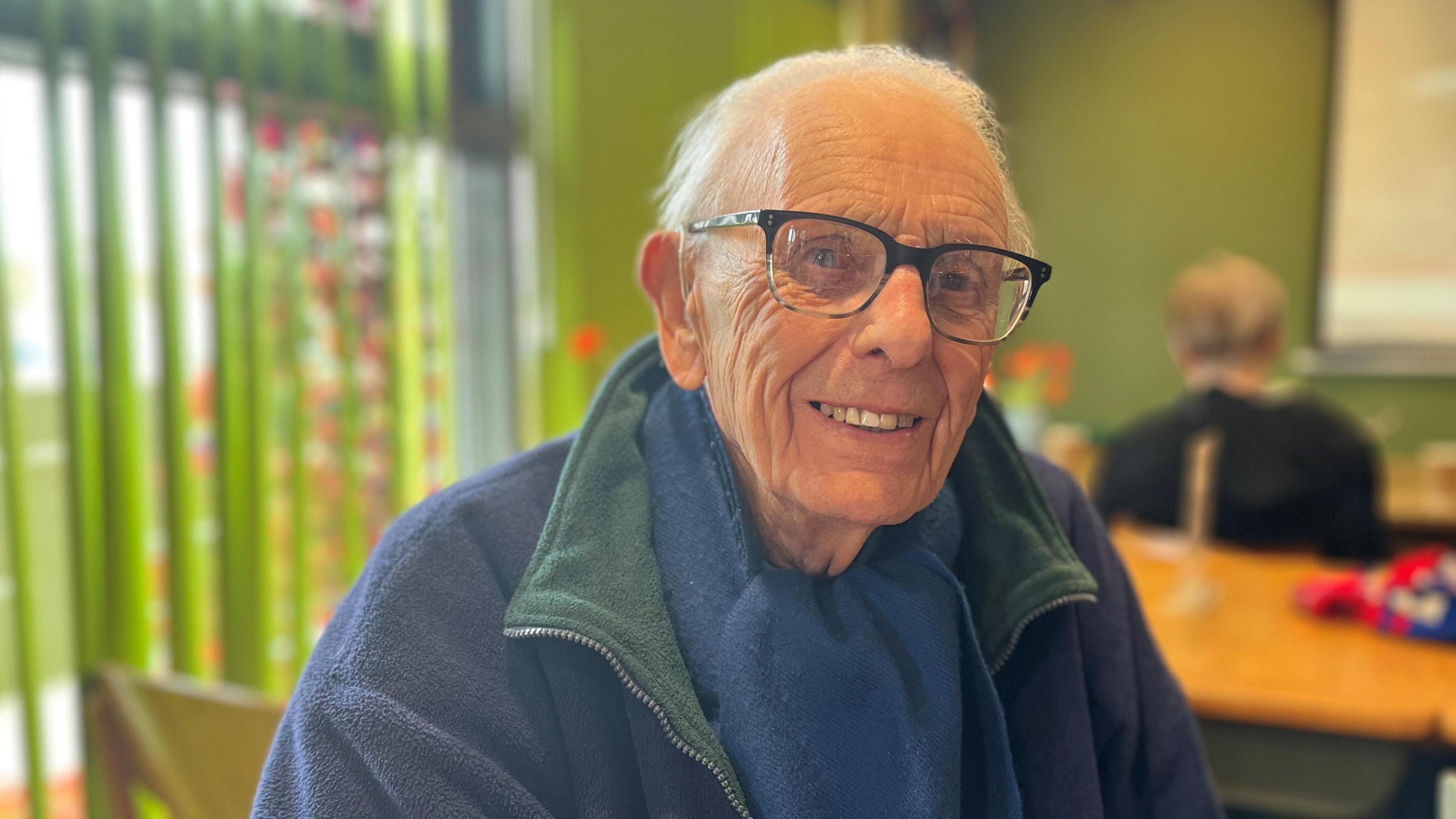 Reg is smiling for the camera. he is wearing a blue fleece top and scarf and has horn rimmed glasses.