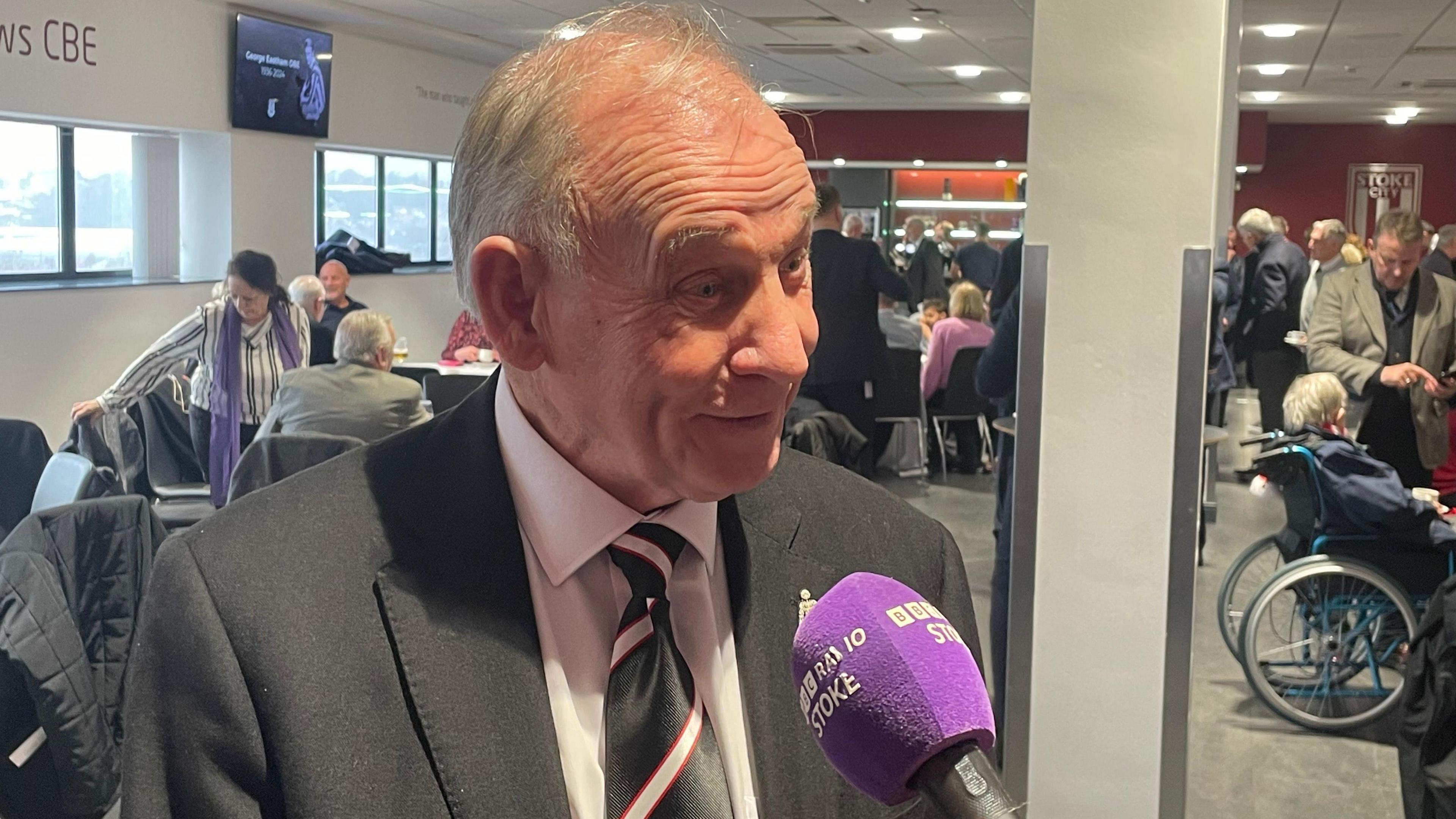 An elderly man being interviewed in a lounge full of people. The man is wearing a black suit jacket, white shirt and black tie with white stripes, with a red outline. He is speaking into a purple microphone that says "BBC Radio Stoke".