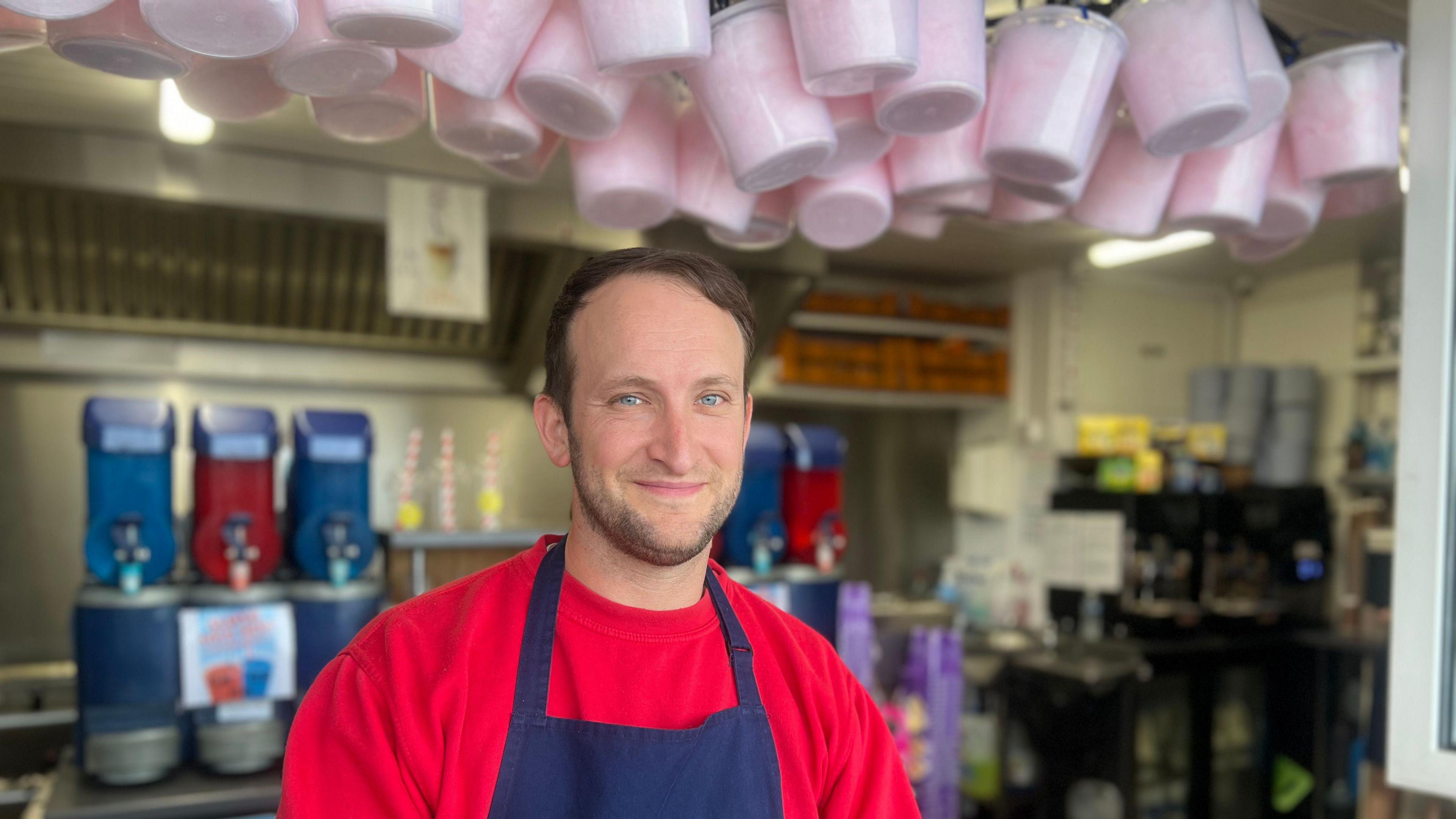 Man in red jumper with dark blue aprons. There are slush machines behind him and candy floss buckets overhead.