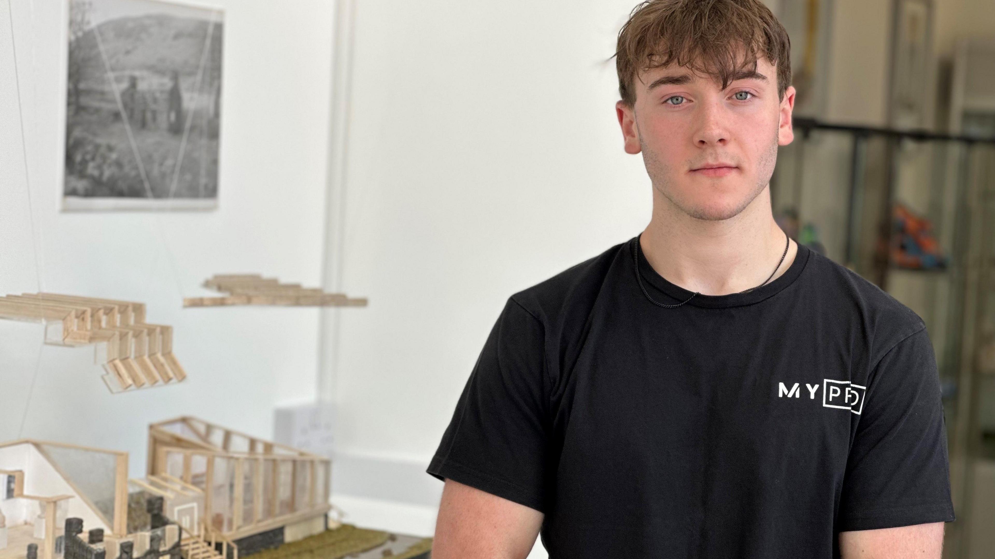 Tom Darnill has short brown hair and is wearing a black T-shirt standing next to a model of a renovated tholtan.