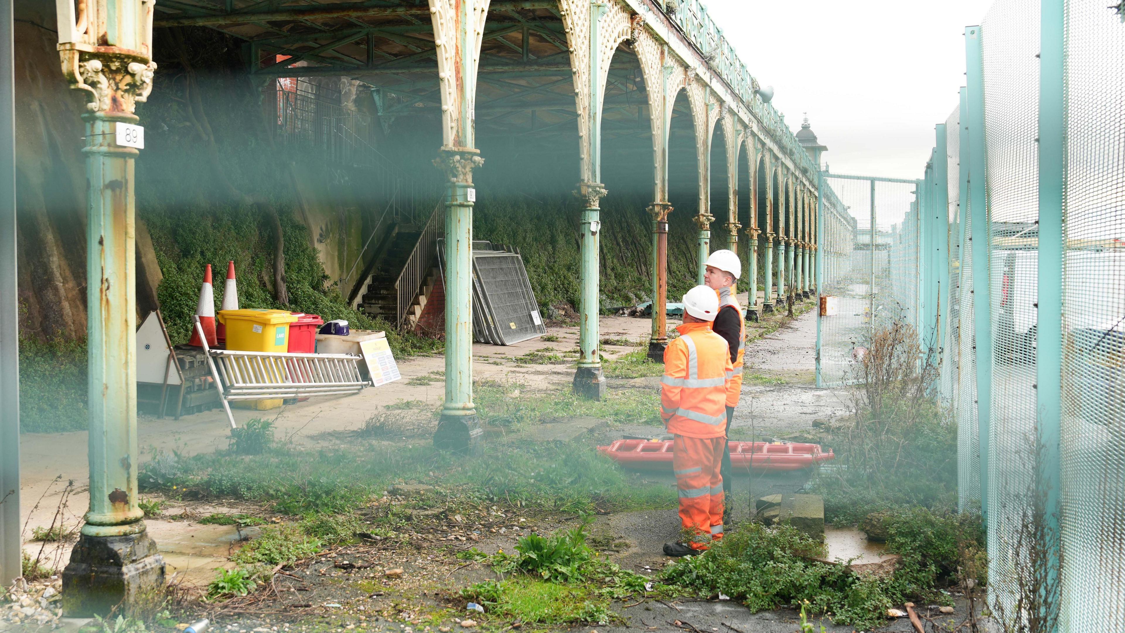 A close up shot between railings of two construction workers in white hard hats and orange high visibility clothing looking a row of historic arches at Madeira Terrace surrounded by debris and weeds