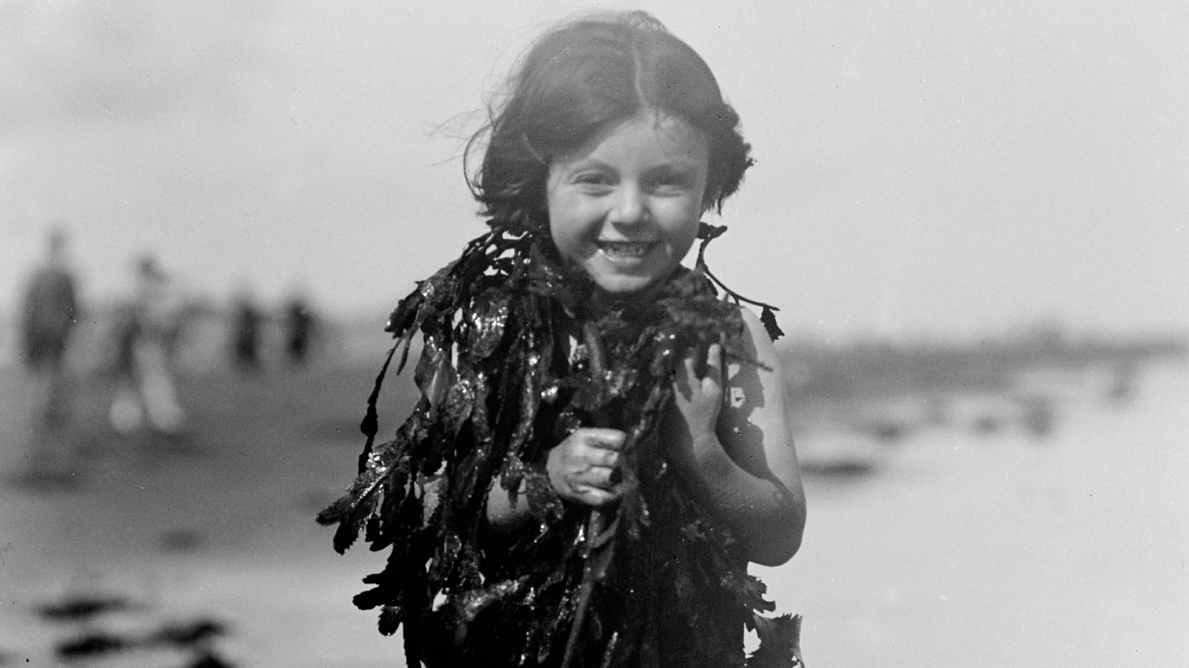 A black and white photos of a young girl at the beach covered in algae. She has dark hair and is laughing at the camera.