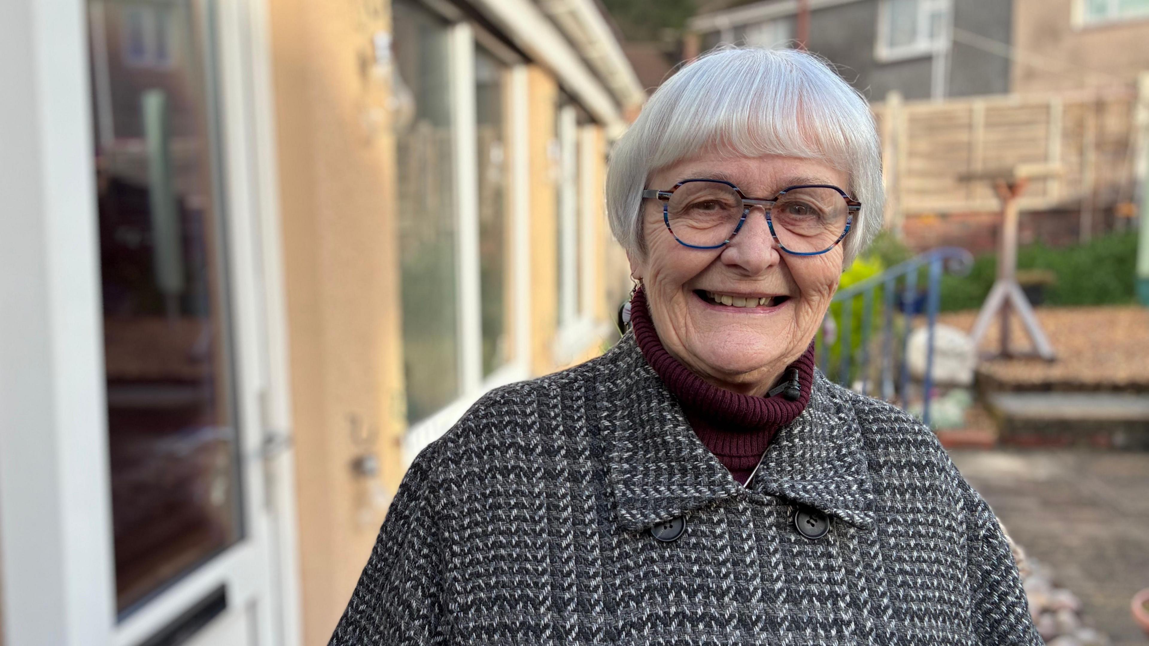 Gail Sydenham yn ei gardd chefn ar b'nawn gaeafol yn gwisgo cot lwyd smart. Mae ganddi wallt gwyn lawr i'w chlustiau ac yn gwisgo sbectolau. yn y cefndir mae rhan o'i chartref gyda fframiau drws a fenestri gwyn, fyddai'n ddu os na fyddai'n eu glanhau'n ddyddiol, medd hi. 