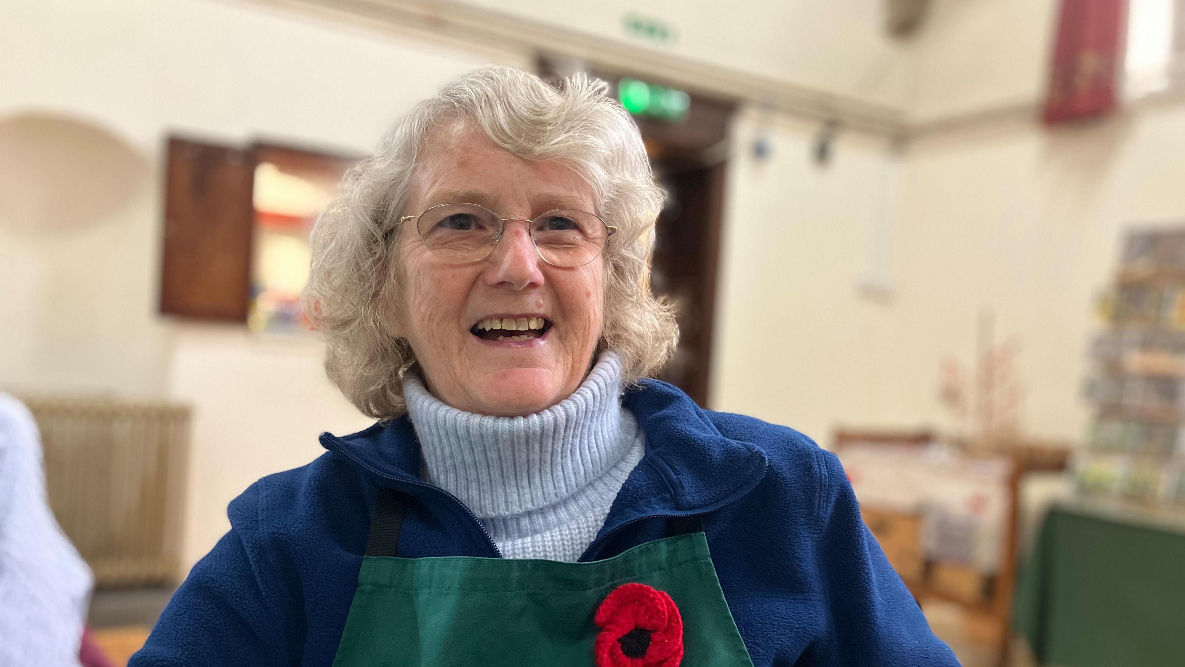 A woman in a light blue roll neck jumper and darker blue jacket with a green apron with a knitted poppy pinned on it sitting in a village hall
