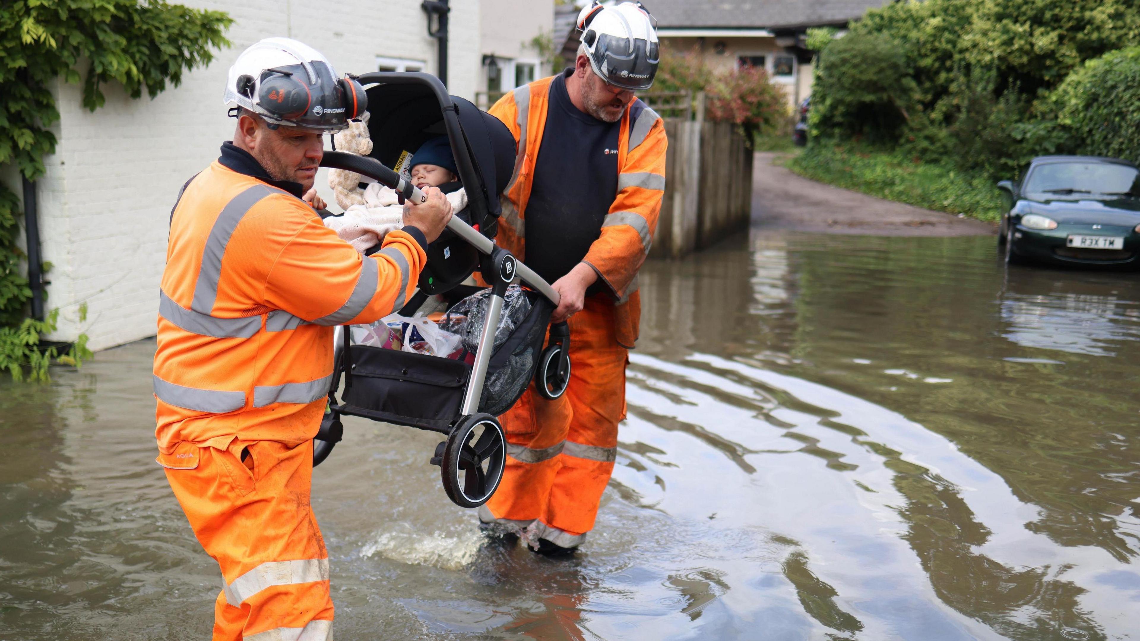 Two men in orange hazard suits and hard hats carry a buggy with a sleeping baby through flood water on a residential street