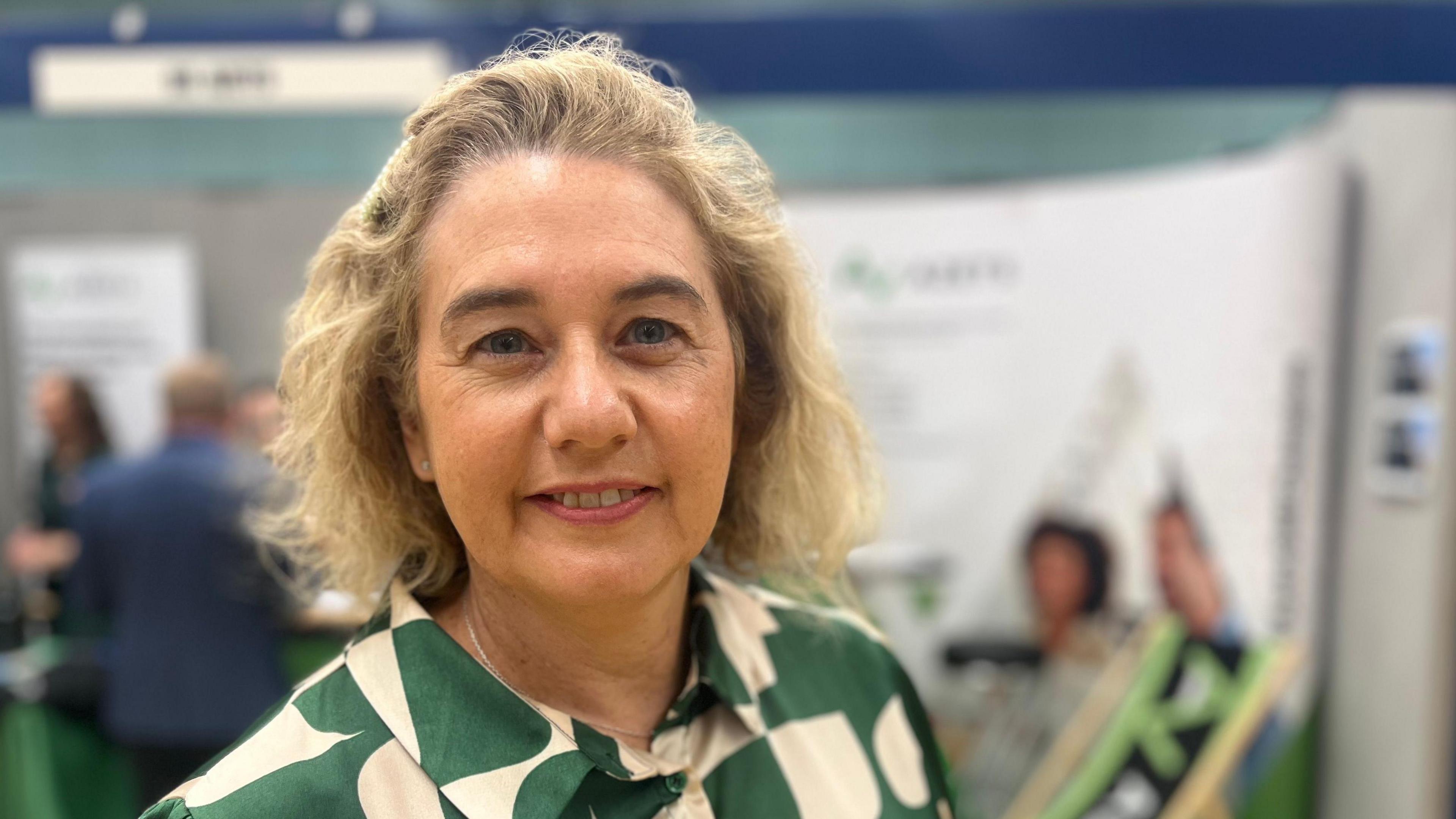 Annette Stone wearing a green and cream patterned blouse in front of a blurred background