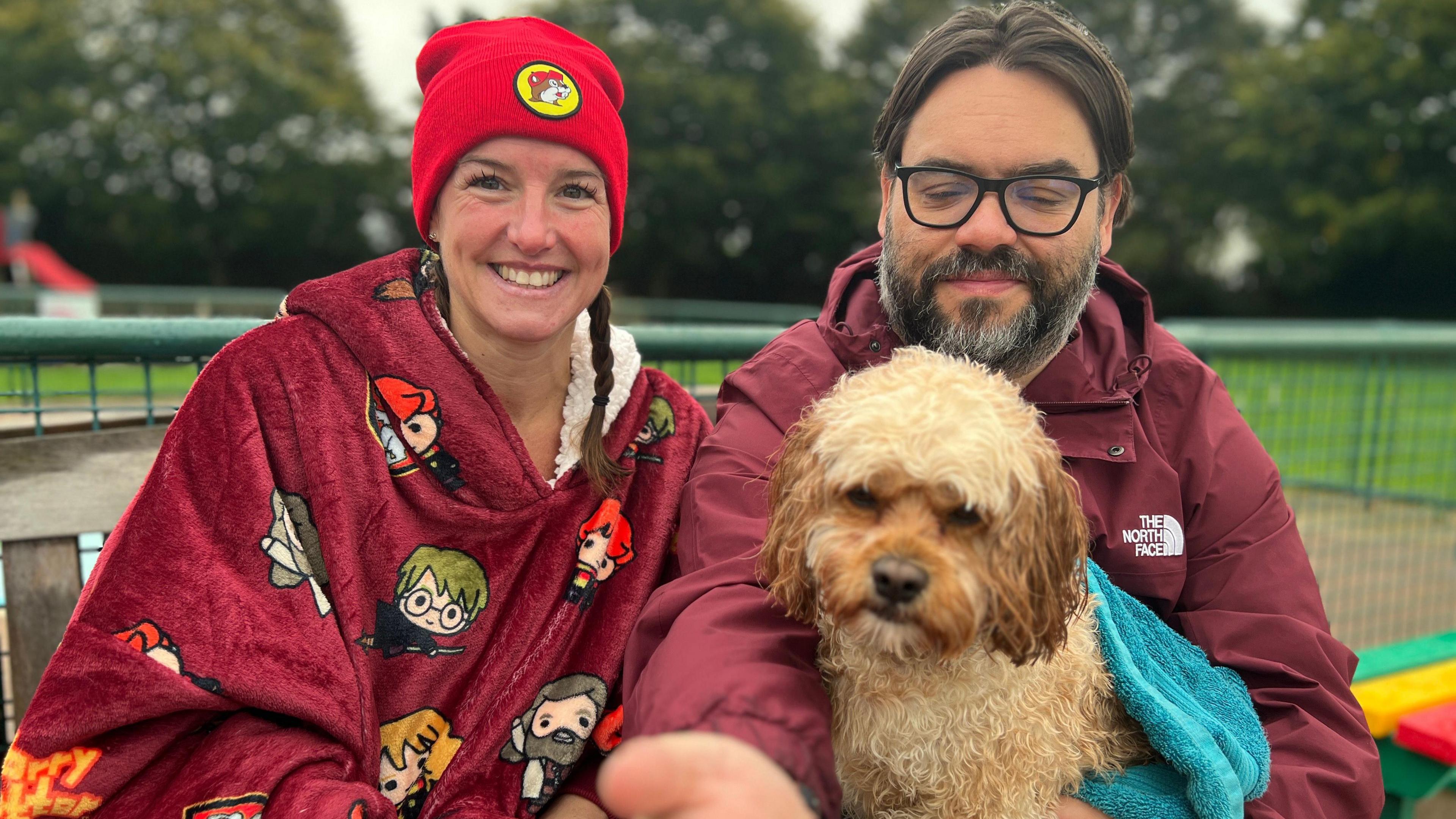 Laura Roe, smiling and wearing a red beanie hat, with a maroon-coloured towel around her, sitting next to a man holding a beige dog on his lap