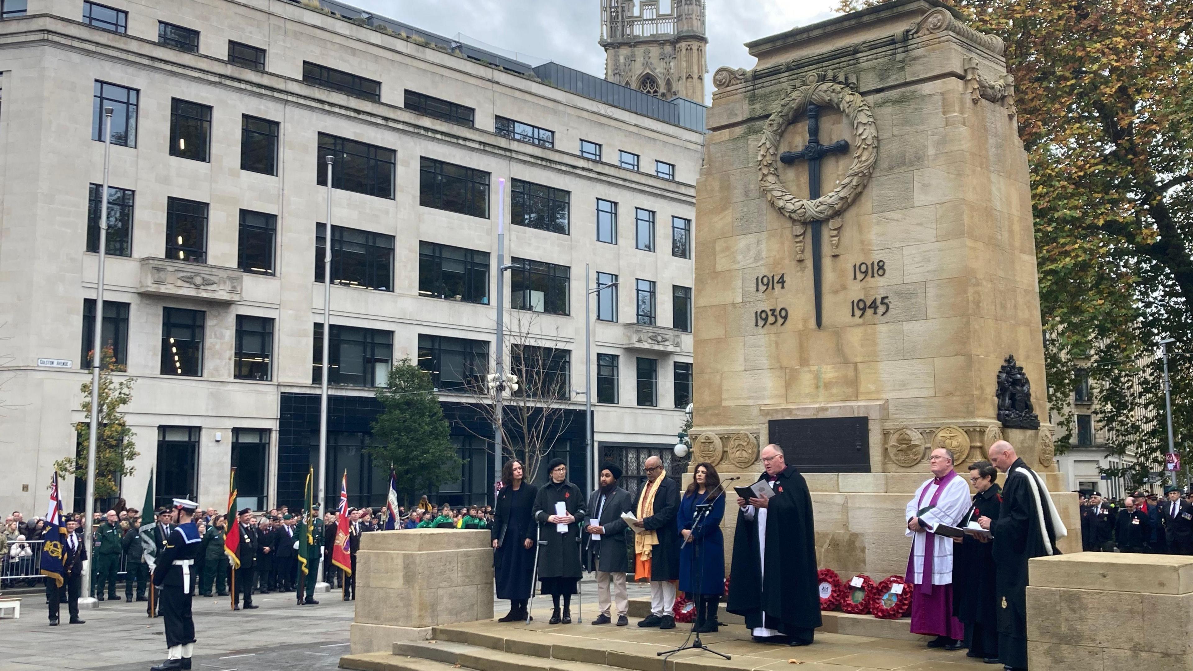 A service at Bristol Cenotaph. There are people standing directly in front of the memorial, speaking into a microphone. Crowds can be seen watching the service.