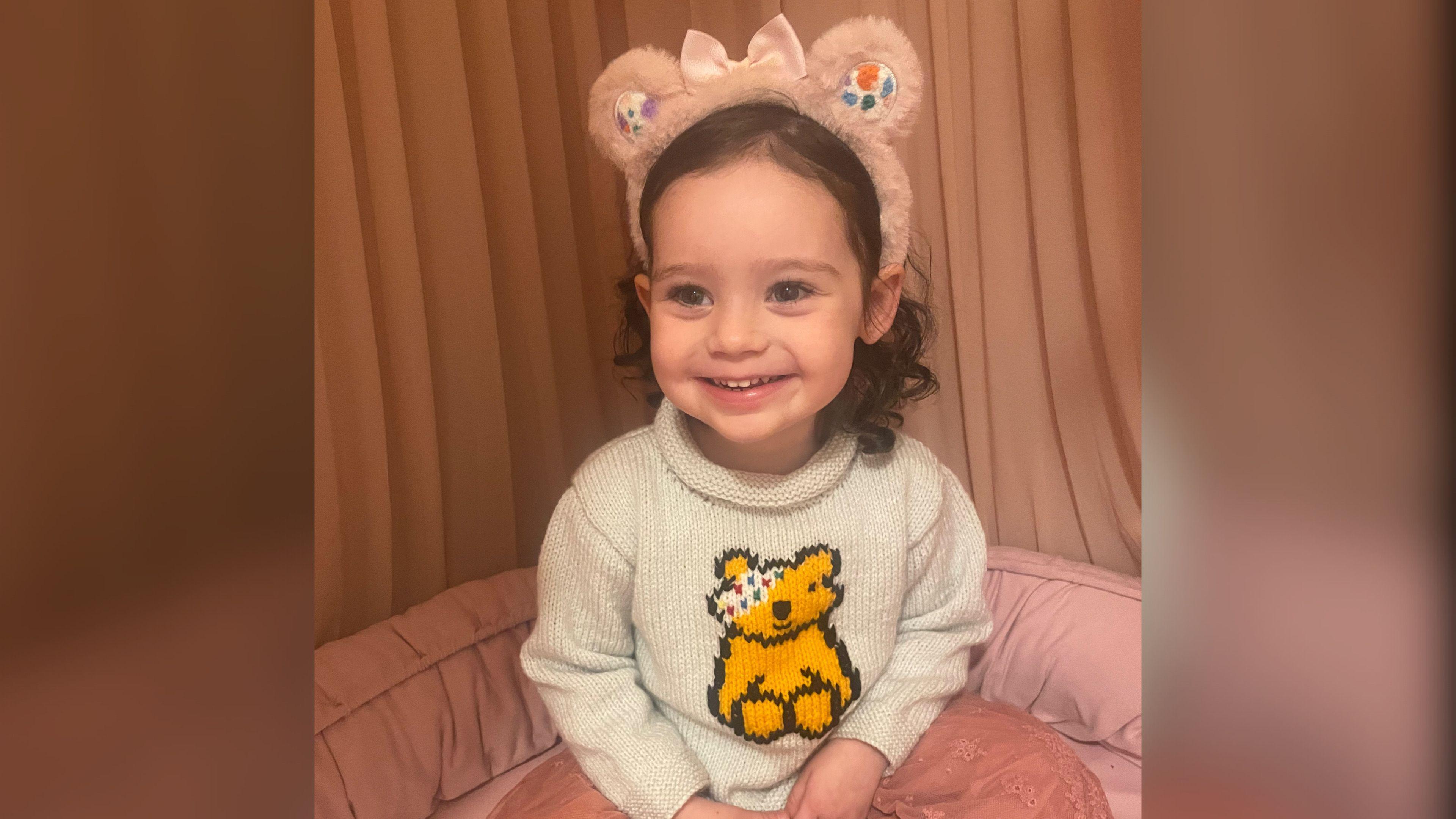 A toddler smiling at the camera wearing a beige jumper with Pudsey on the front