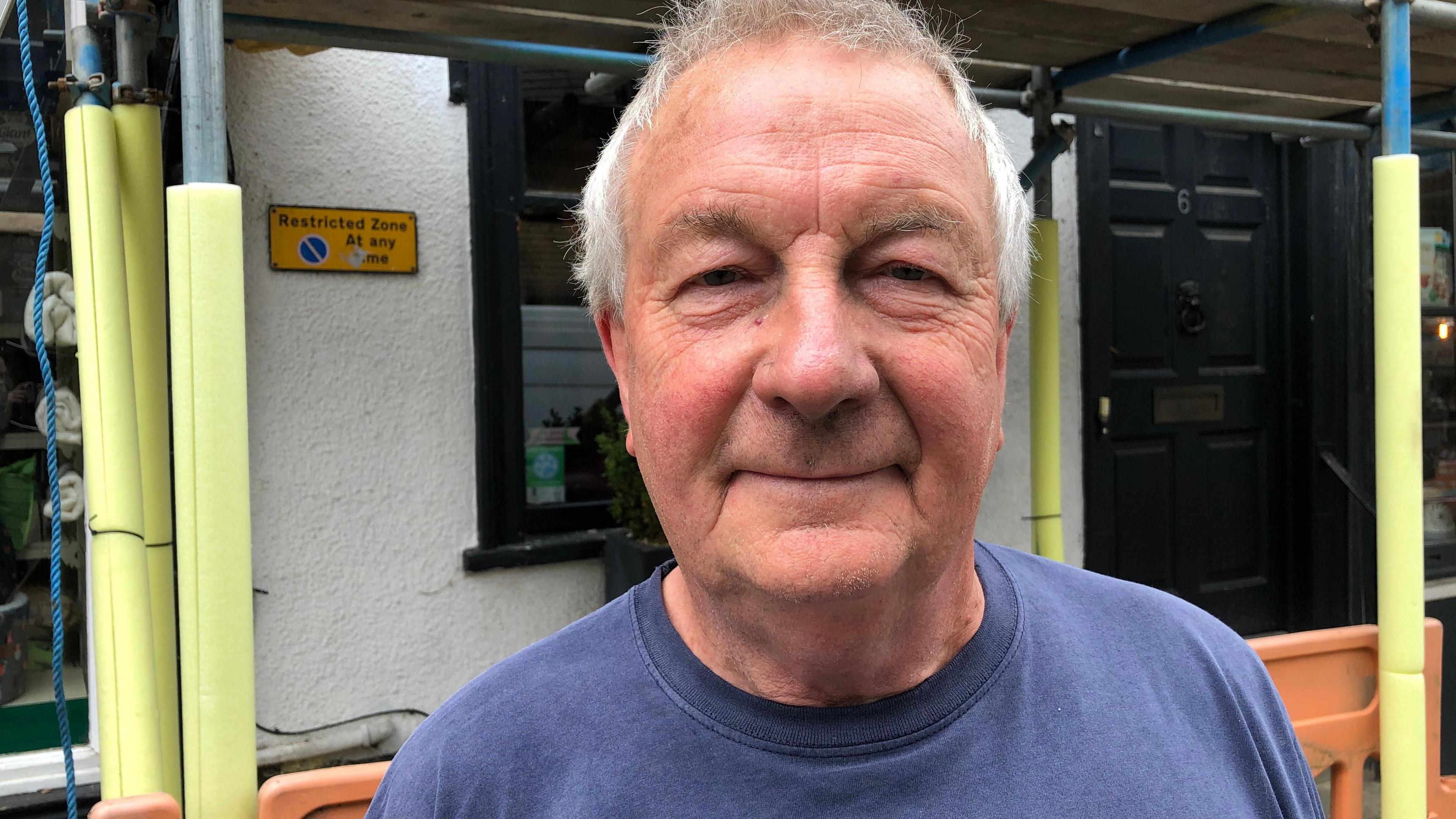 A man in a mid blue sweatshirt standing in front of a building covered in scaffolding