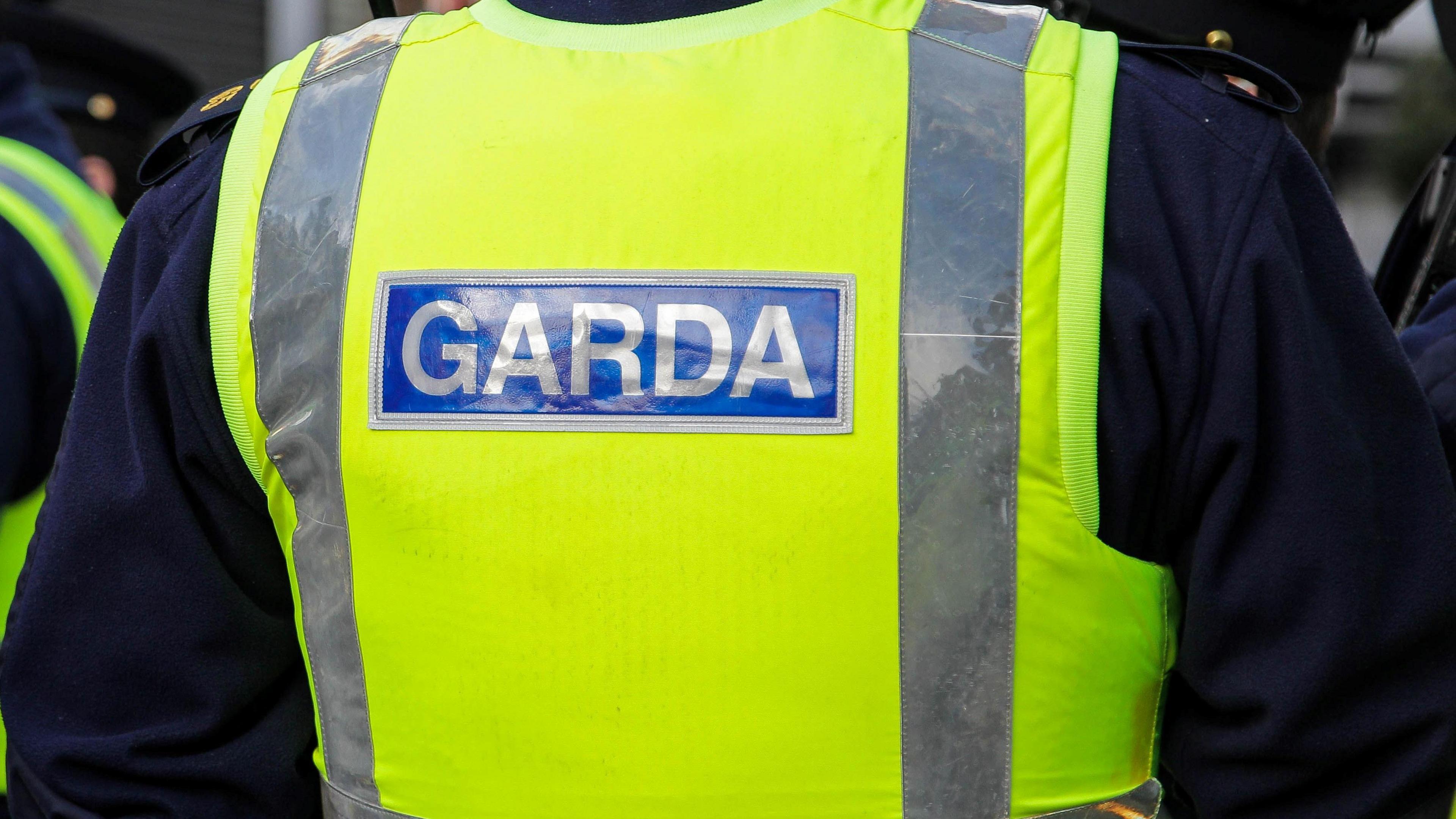 A garda's back. He is wearing a yellow high vis jacket with garda printed across it. Some other officers are standing close to him.