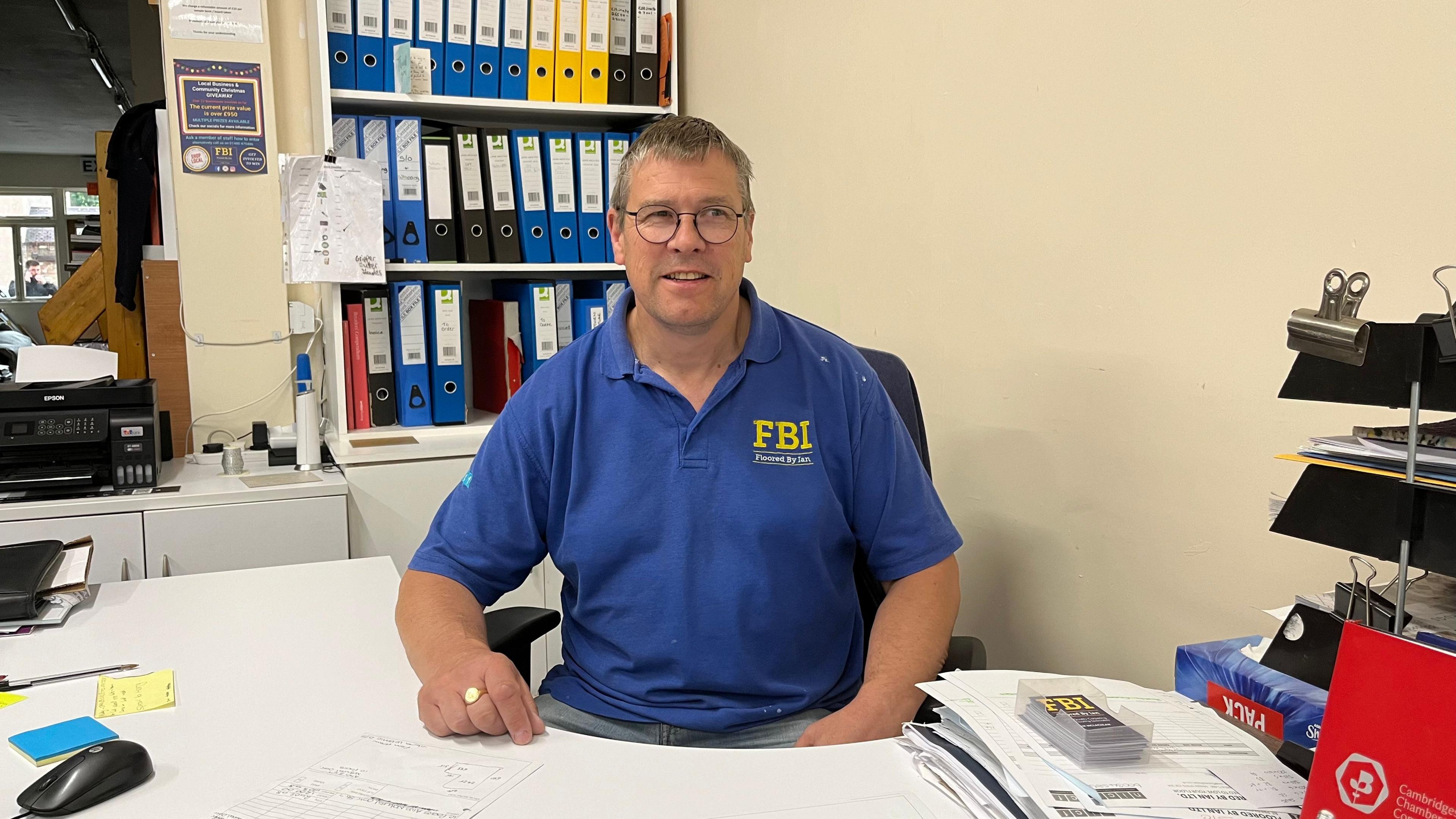 Ian McLachlan sat at a white desk with blue, red, black and yellow filed behind him. He wears a blue shirt with the yellow FBI (Floored by Ian) logo in the top right. He has one hand on the desk, a ring on his finger and wears circle framed glasses. 