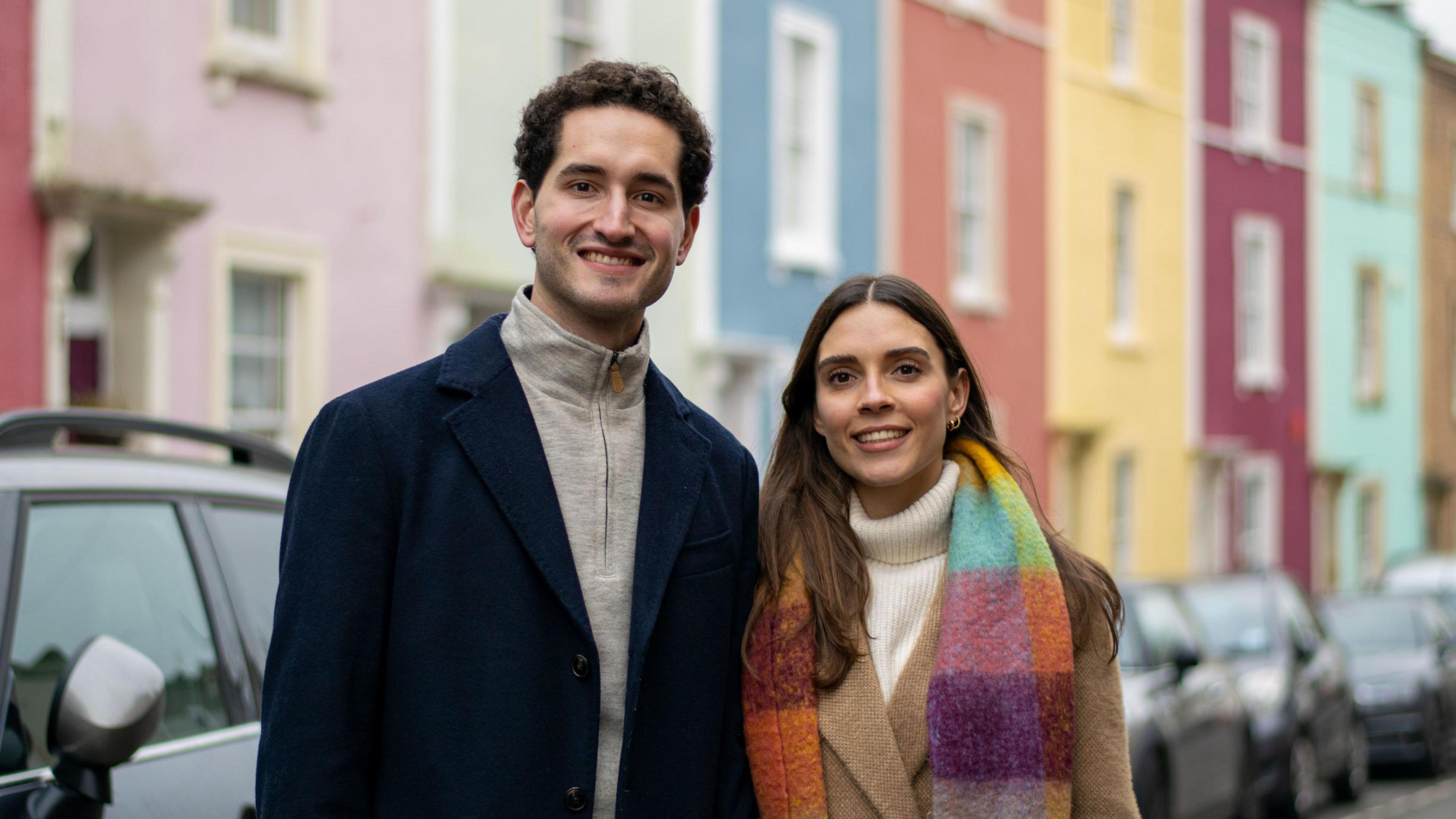 A man and a woman stand on a street in Bristol with colourful houses in the background. He is wearing a black long jacket and a grey fleece top. She is wearing a colourful scarf, fawn-coloured jacket and a white rollneck jumper. They are both smiling