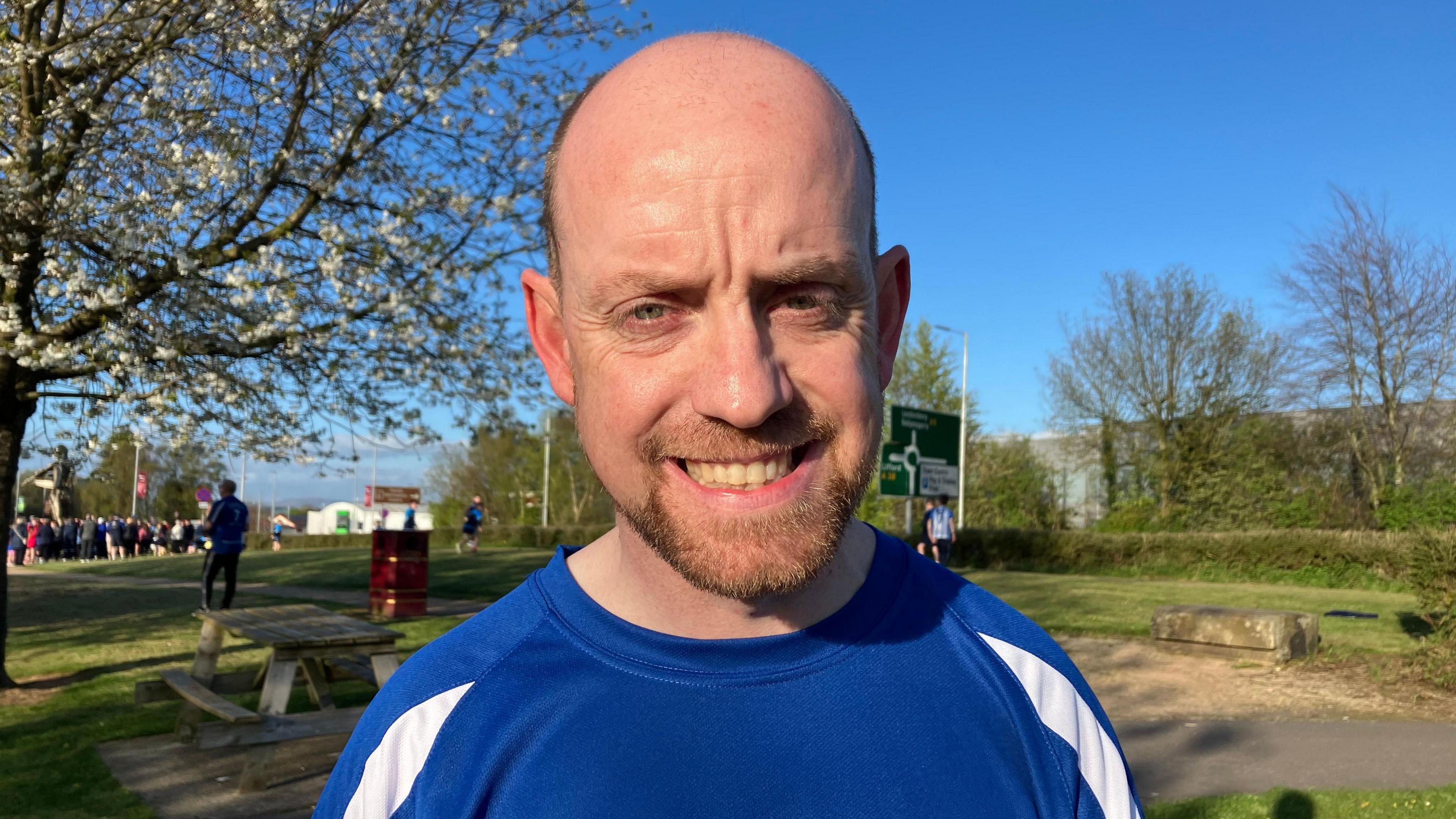 Adrian Loughrey. He is standing in a park with a picnic table and a tree to the left. He is partly bald and wearing a blue and white t-shirt.