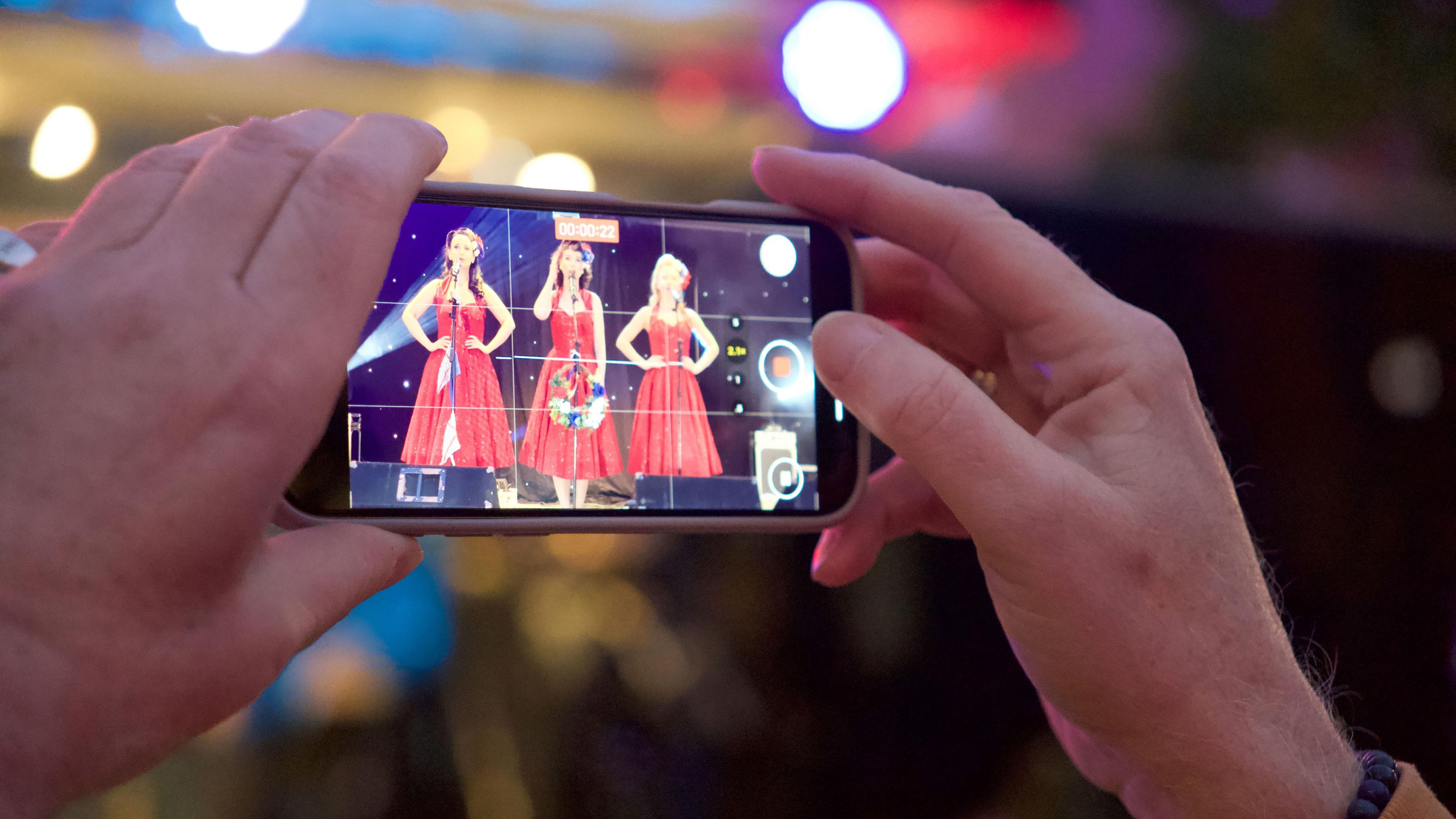 A man holding up a phone to record three singers in red dresses