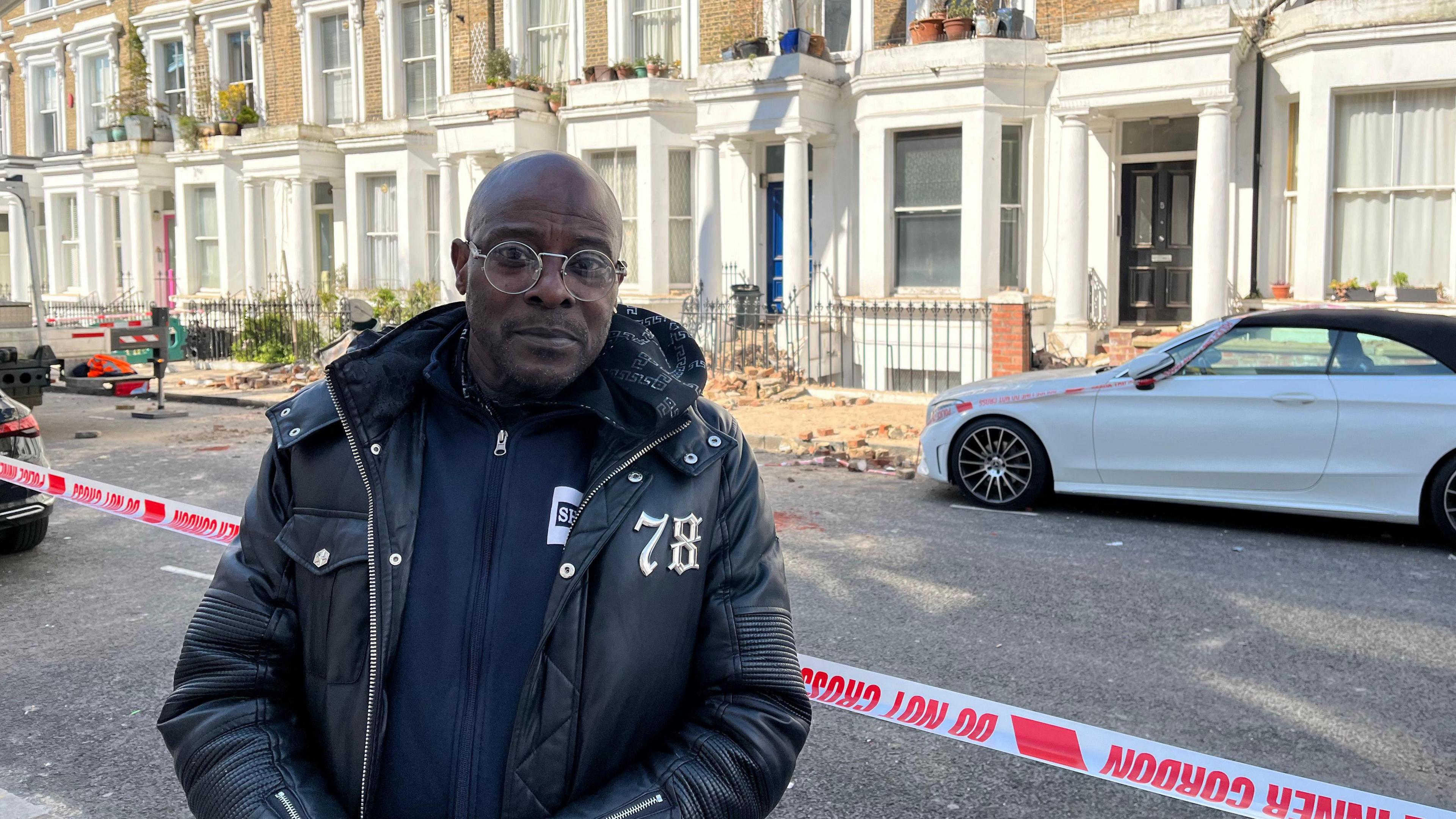 John Bedeau is looking into the camera in front of 'do not cross' tape, with rubble and bricks in the background. He has glasses on, wearing a black jacket and blue hoodie. 