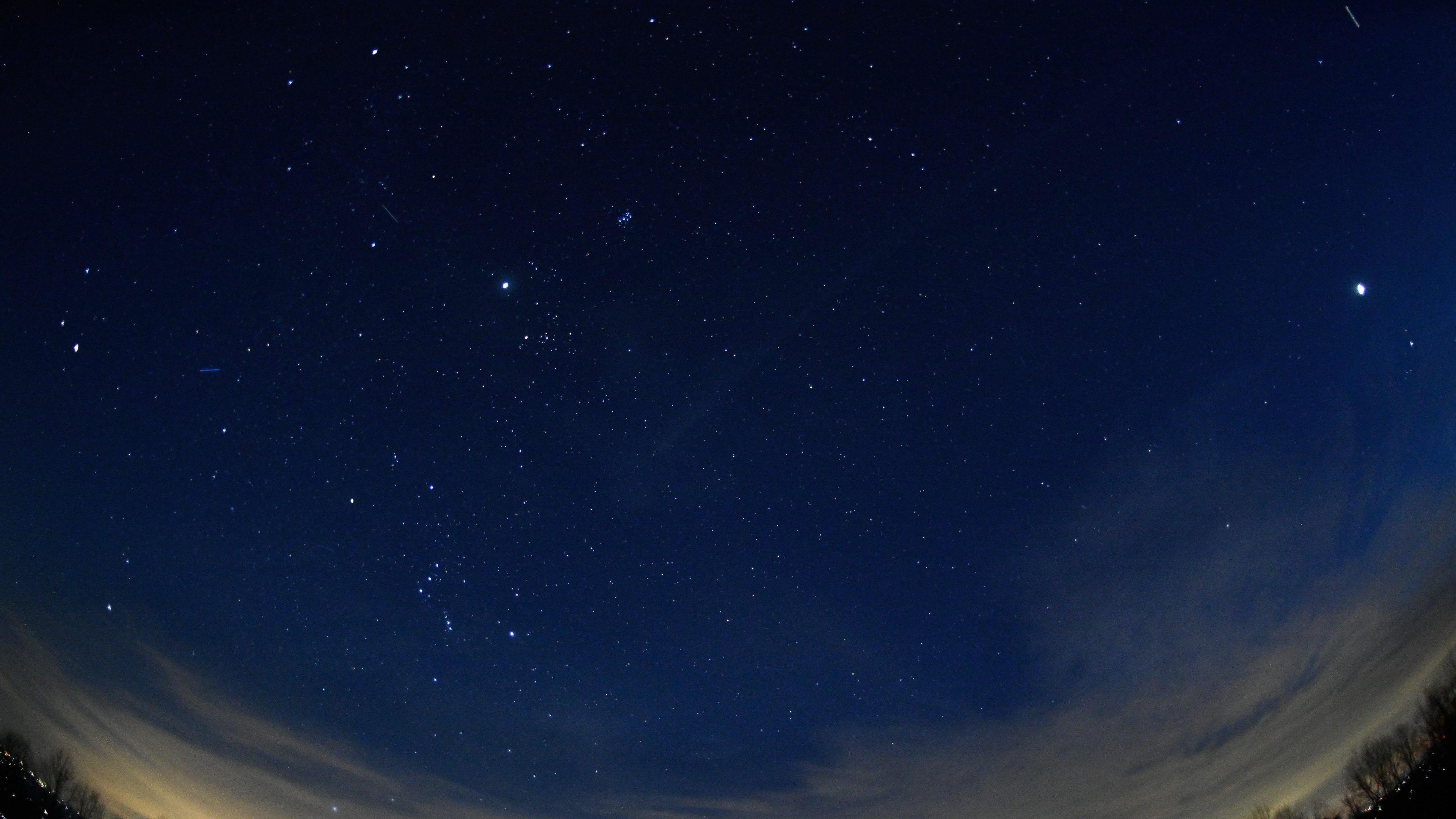 A stunning wide-angle nighttime photograph of the evening sky, featuring four planets. 

Mars on the left, Jupiter in the middle, and Saturn and Venus on the right. Wispy clouds stretch across the lower part of the sky, with the silhouettes of trees and city lights glowing faintly. 