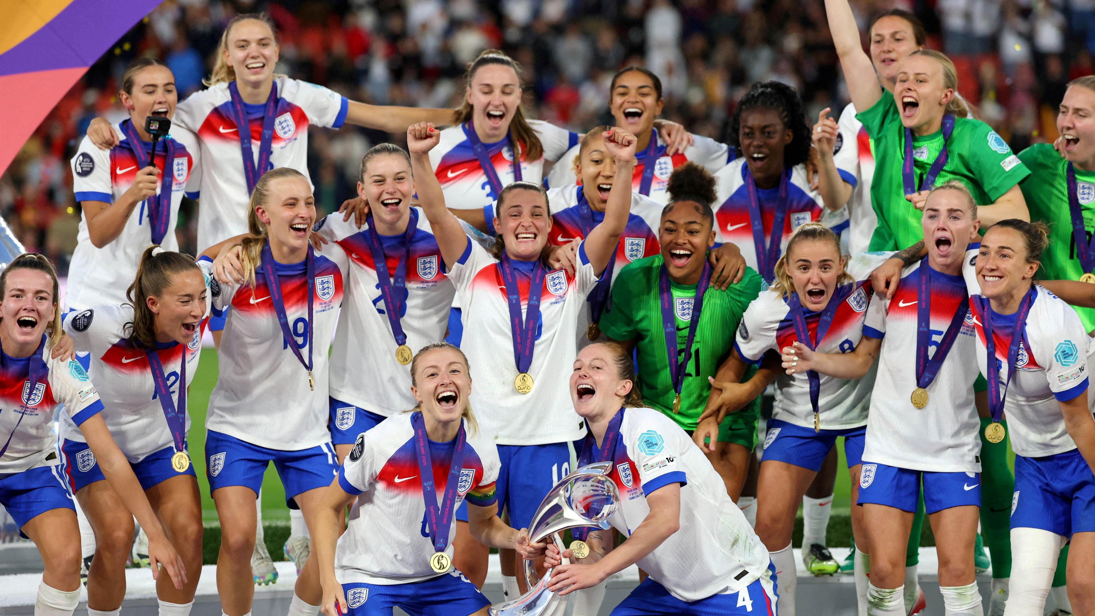 England's Leah Williamson and Keira Walsh lift the trophy as they celebrate with teammates after winning the UEFA Women's Euro 2025. All the players are in their white kit with blue shorts and have medals around their necks. They are cheering and smiling.