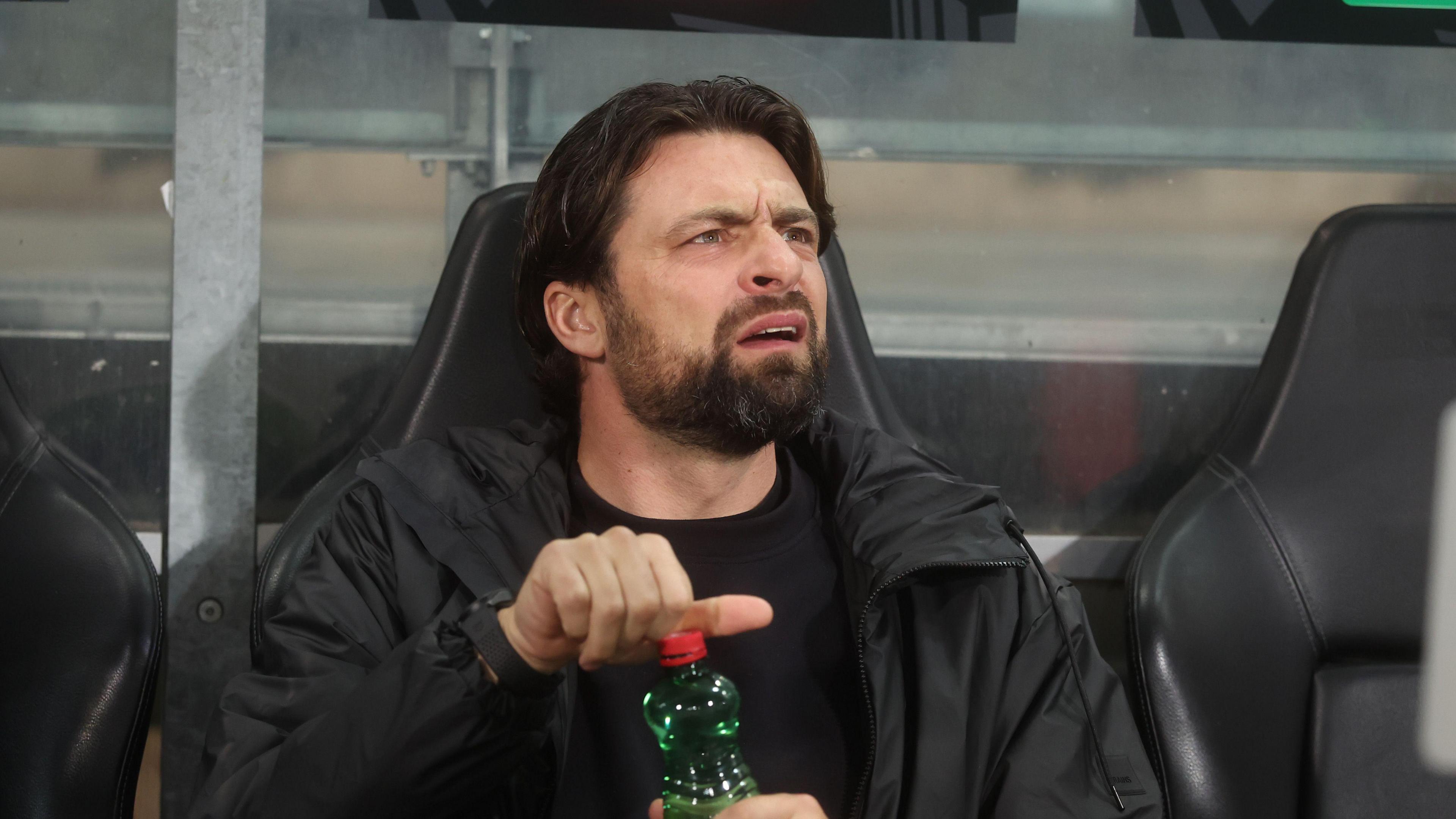 Russell Martin, sitting in the visitors' dugout at SK Sturm Graz while managing Rangers in 2025, looks confused as he opens a green plastic bottle with a red lid