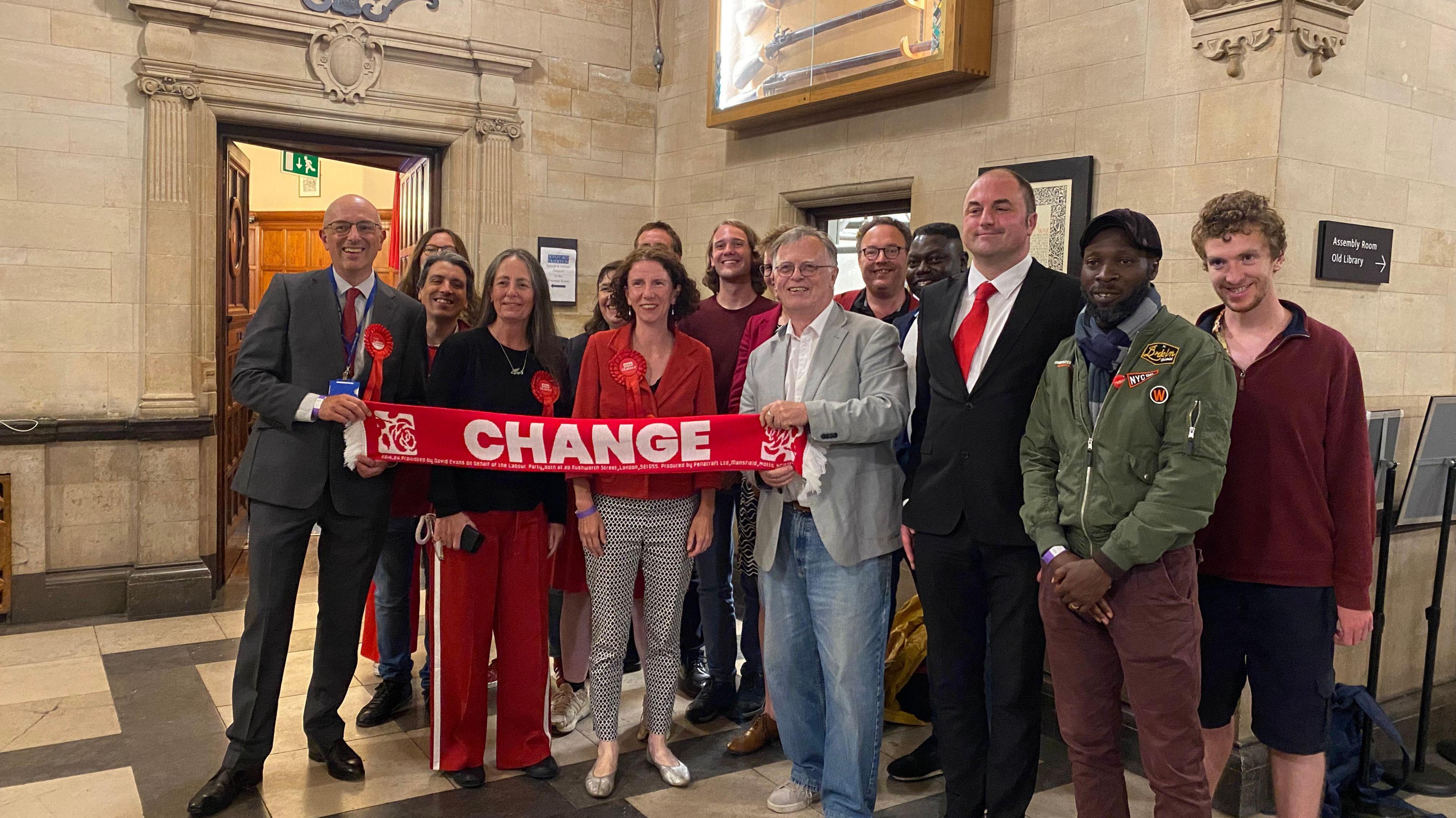 Anneliese Dodds wearing a red Labour rosette behind a Labour scarf with "change" written on it surrounded by Labour supporters inside Oxford Town Hall