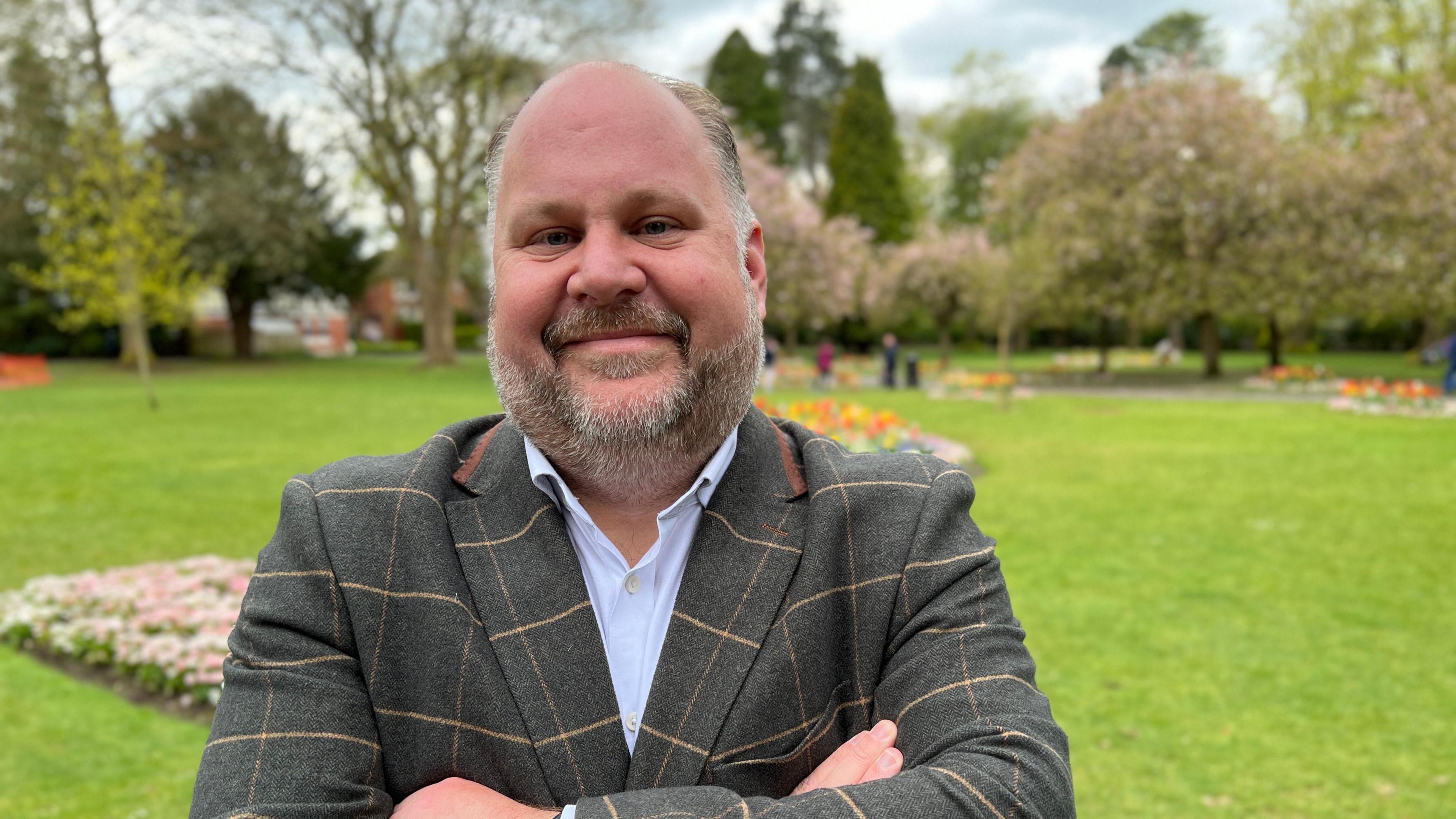 Jim Robbins stands with arms folded, wearing a shirt and blazer in a park