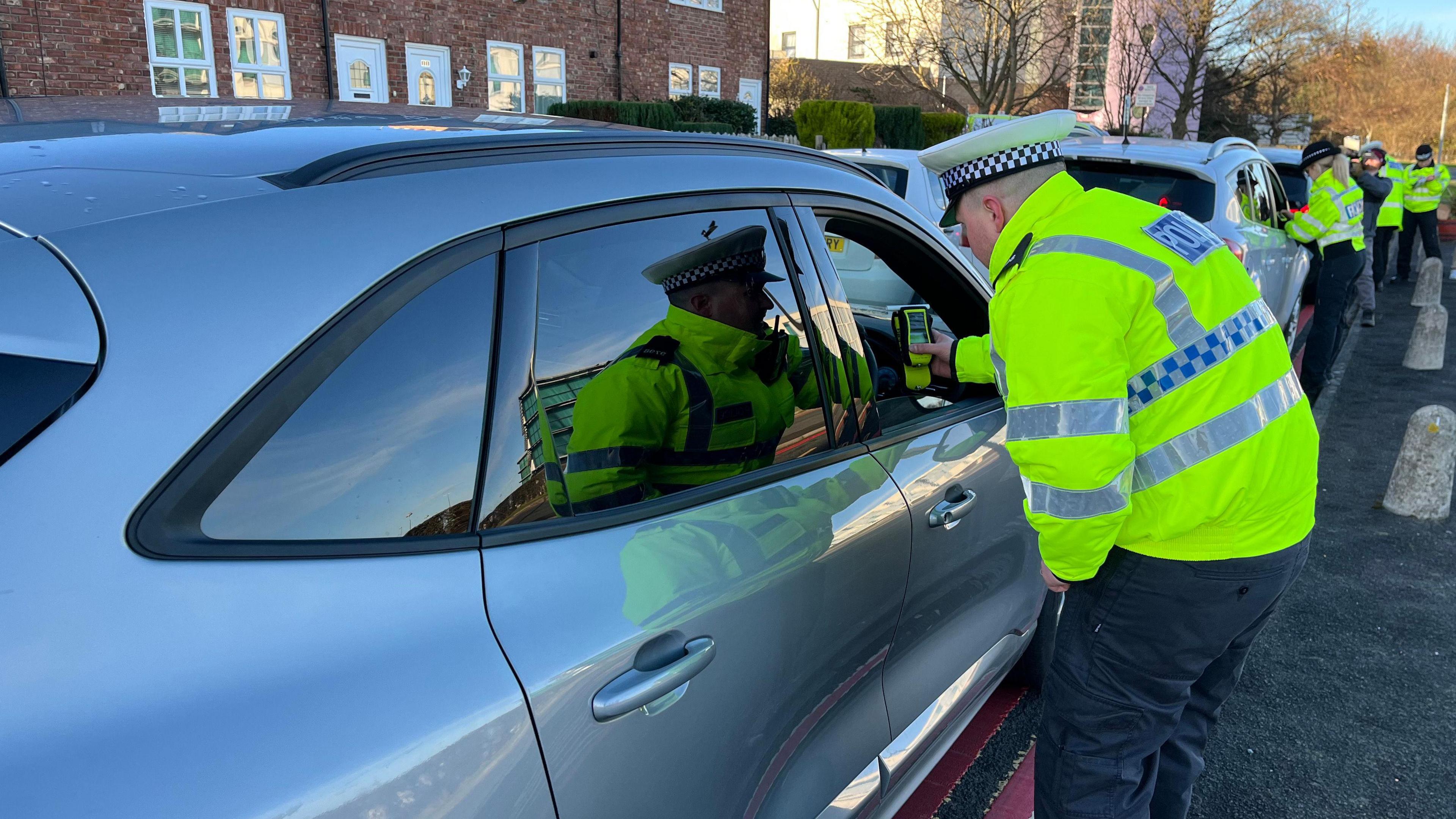 A police officer wearing a hi-vis jacket passes a breathalyser test to a driver sat inside a car. The car is one of a number lining a road. Police officers are standing at each car carrying out similar tests.