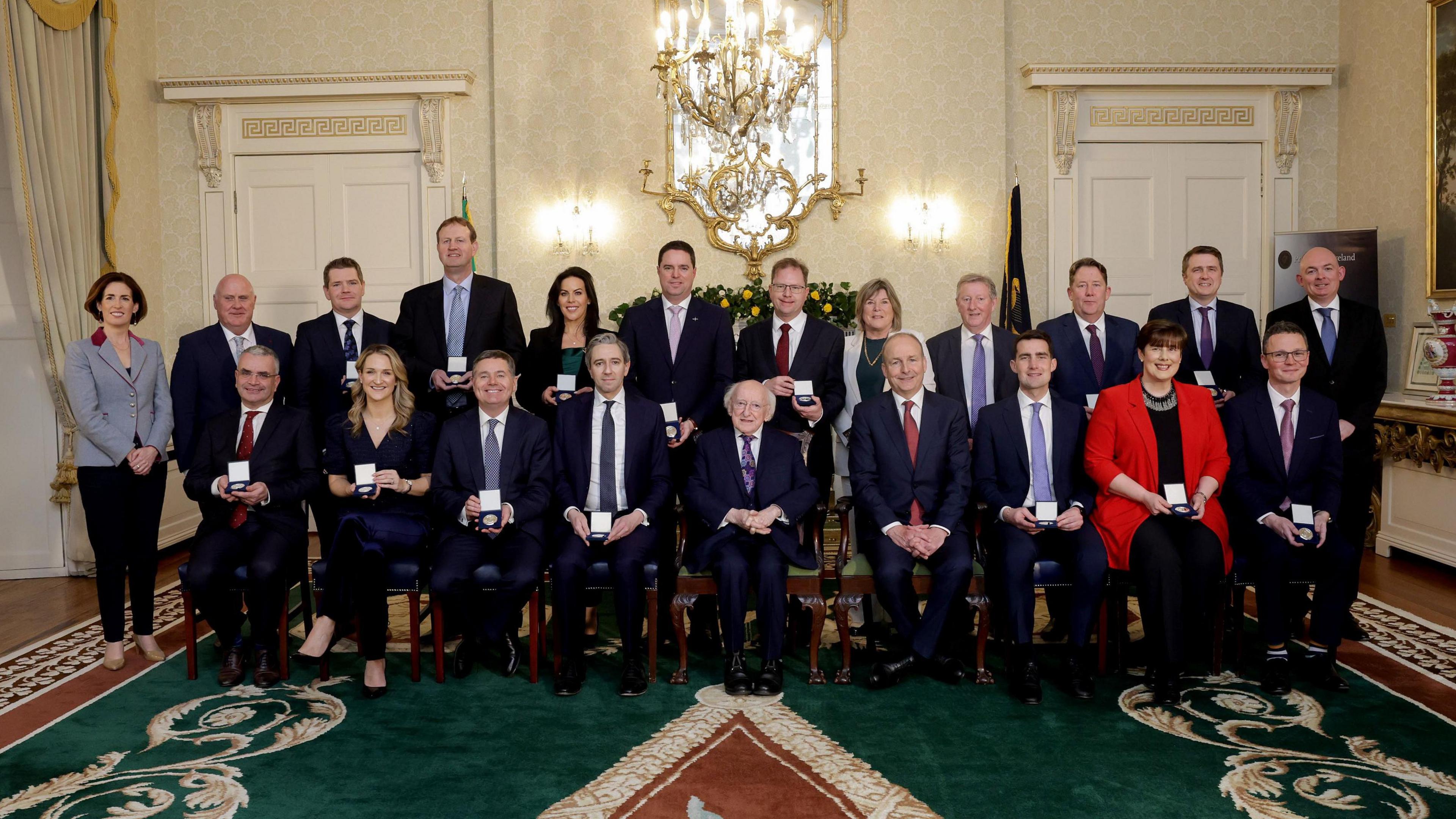 9 people sit in wooden chairs in front of 12 people who stand. All except two hold white boxes in their hands. The carpet beneath them is dark green and the walls are cream. A golden chandelier hangs above them. 