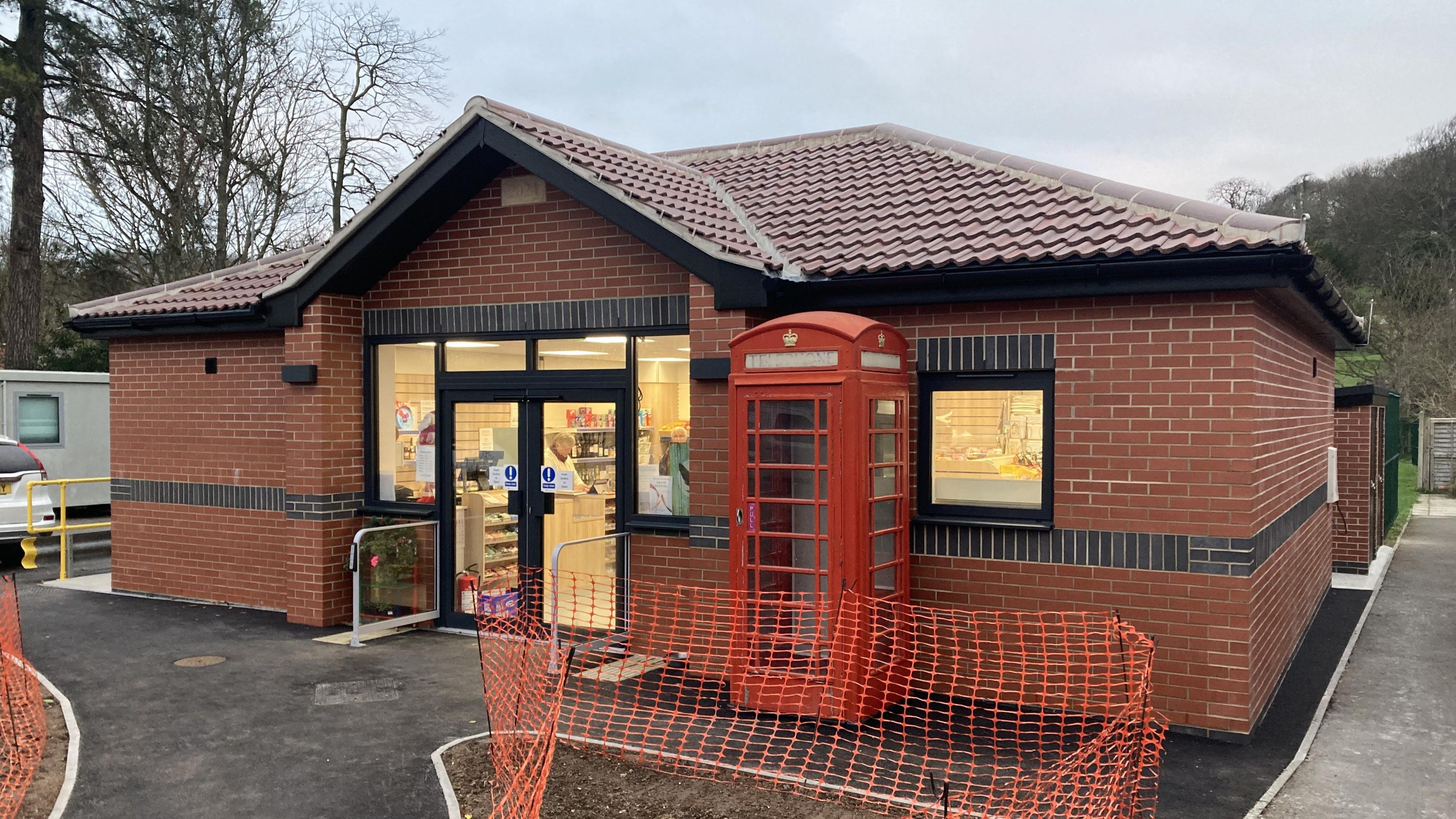 A single-storey brick building in the middle of the shot, with a red phonebox infront of it. There is some orange safety netting on the areas not on the path leading up to the shop. In the background there is a grey sky.