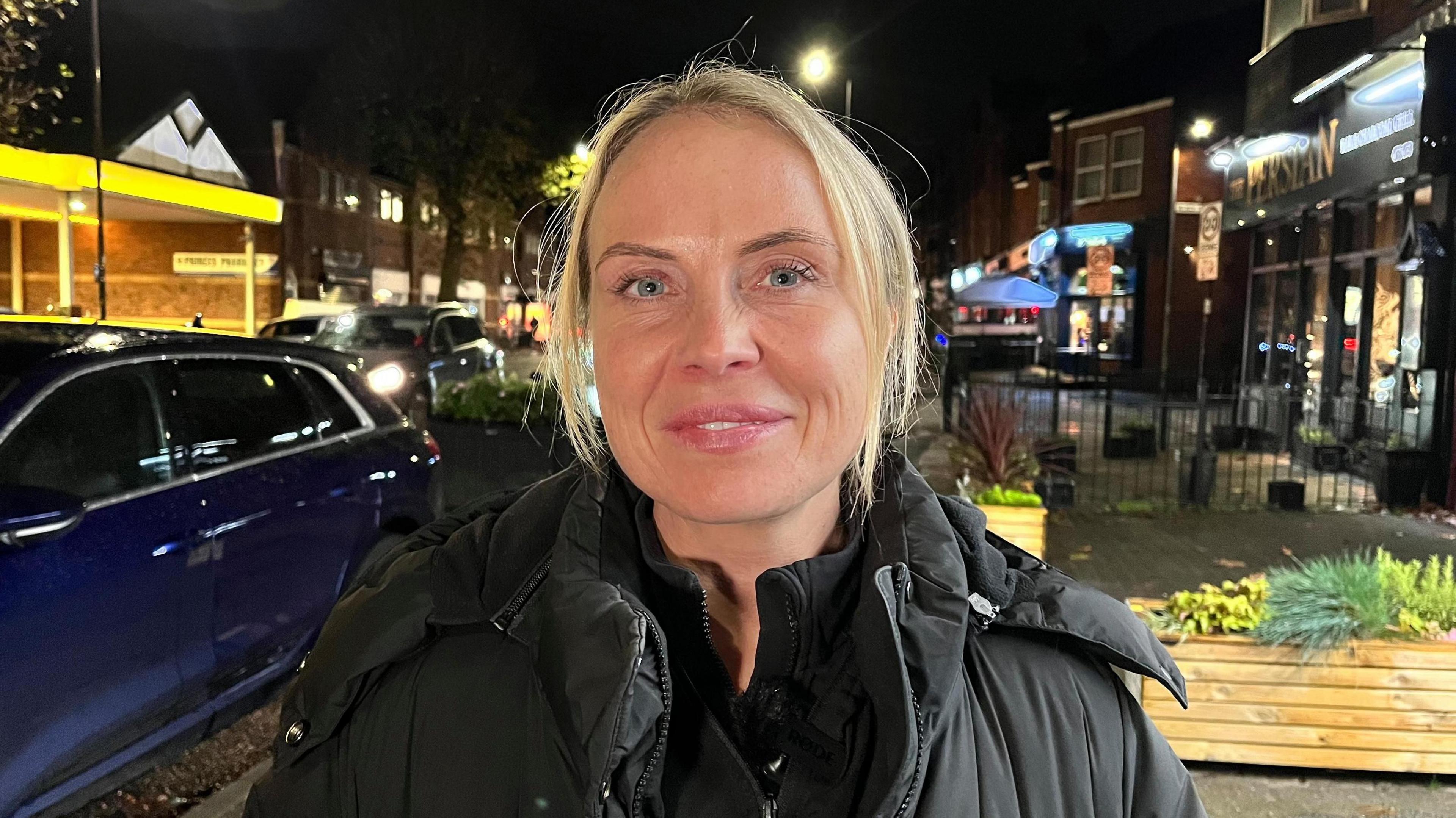 Bev stands in a busy Hull street with bars and restaurants in the background. She is wearing a black coat and smiles at the camera. She has blonde hair parted.