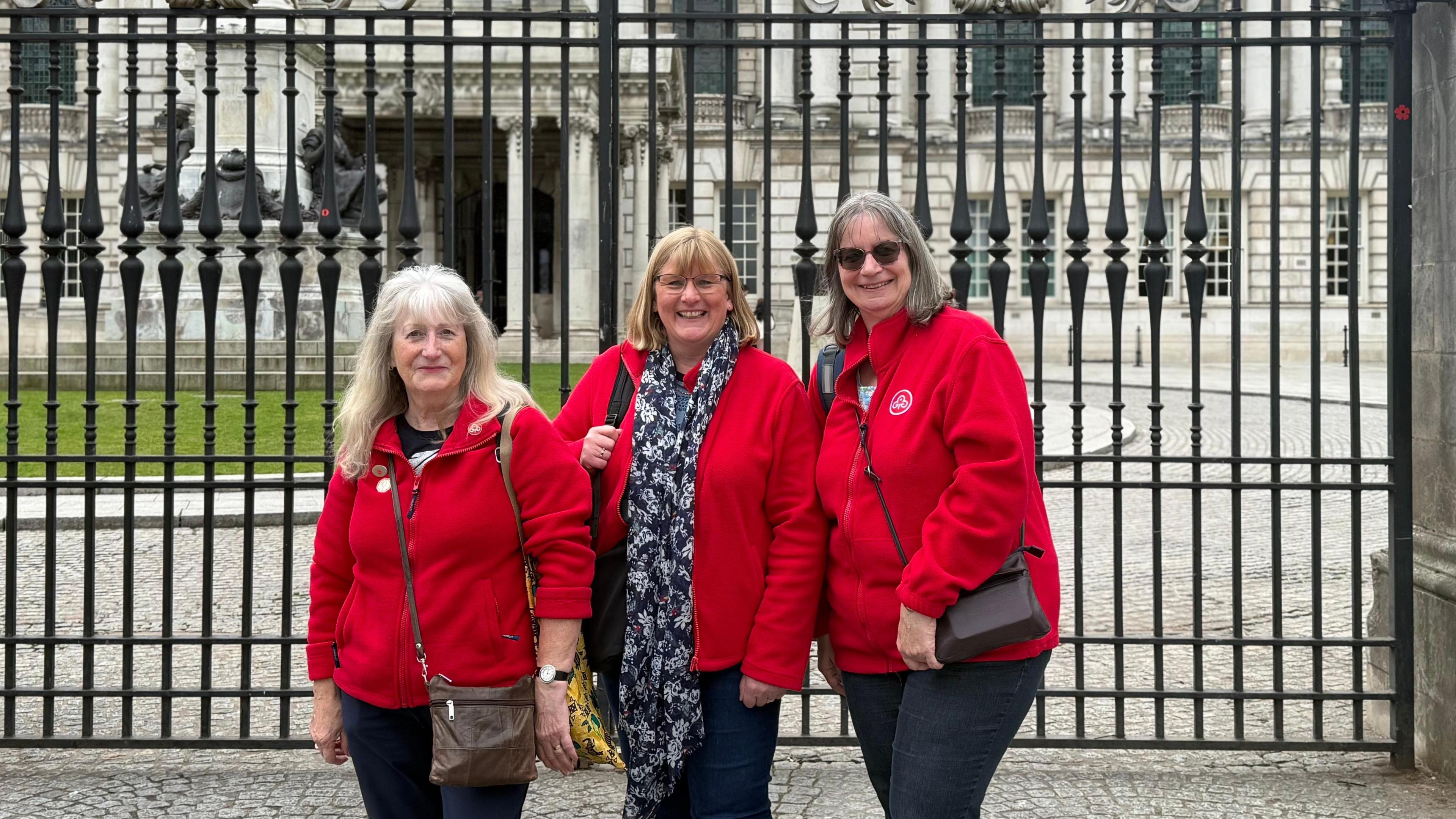 Three ladies in red fleeces with a Trefoil flowery badge stand in front of railings, behind them is a grand looking white washed Belfast City Hall.
The lady on the left has white long hair and a fringe with a brown satchel bag over her red fleece.
The middle lady has short blond hair with a fringe and a yellow bag and flowery blue and white patterned scarf on.
The lady on the right has grey hair, dark sunglasses on and a leather looking brown bag across her red fleece.