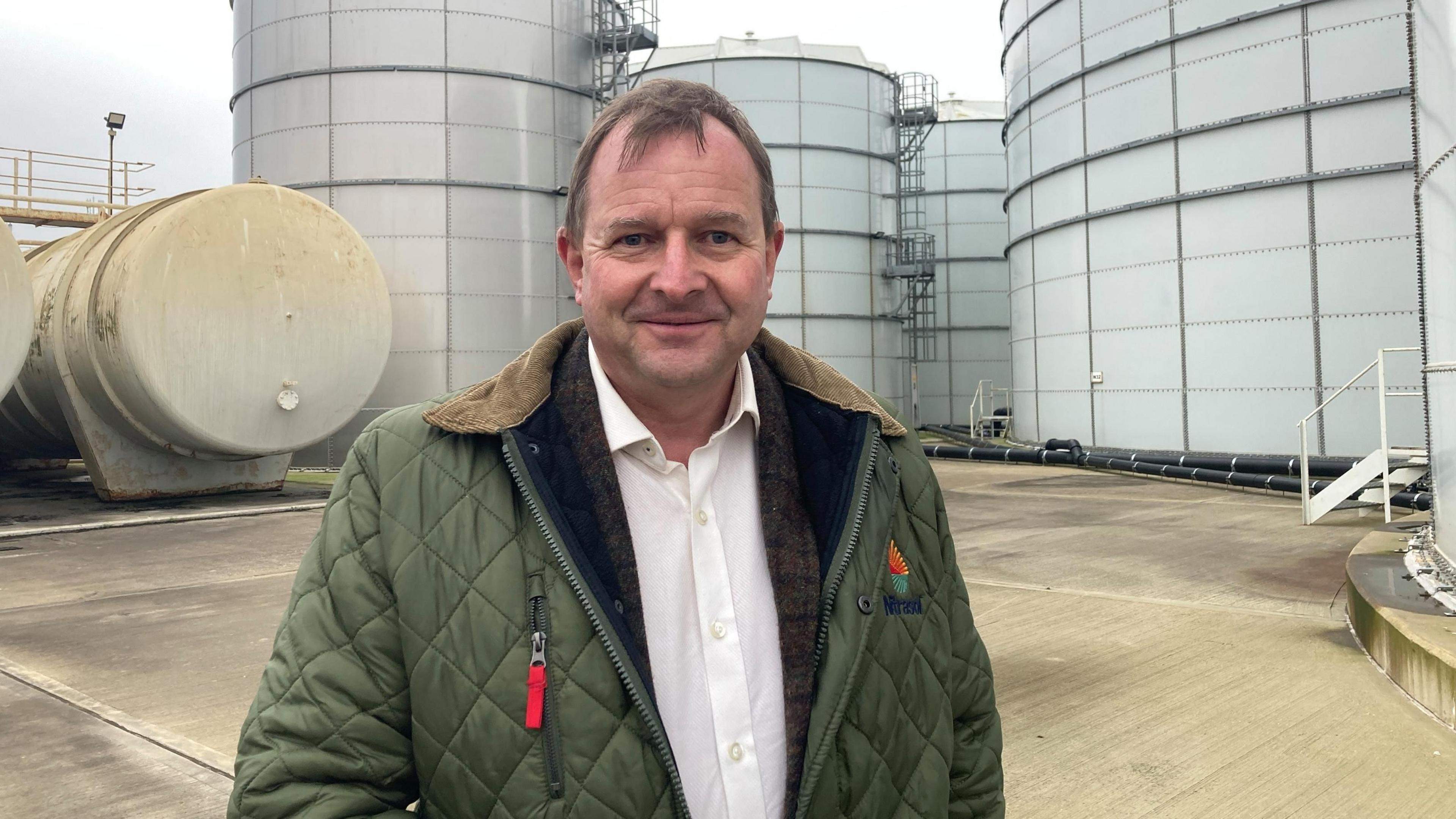 Lord Fuller, former leader of South Norfolk Council, has short, brown hair and is wearing a green coat with a brown jacket underneath and a white shirt.
He is standing in front of large silos.