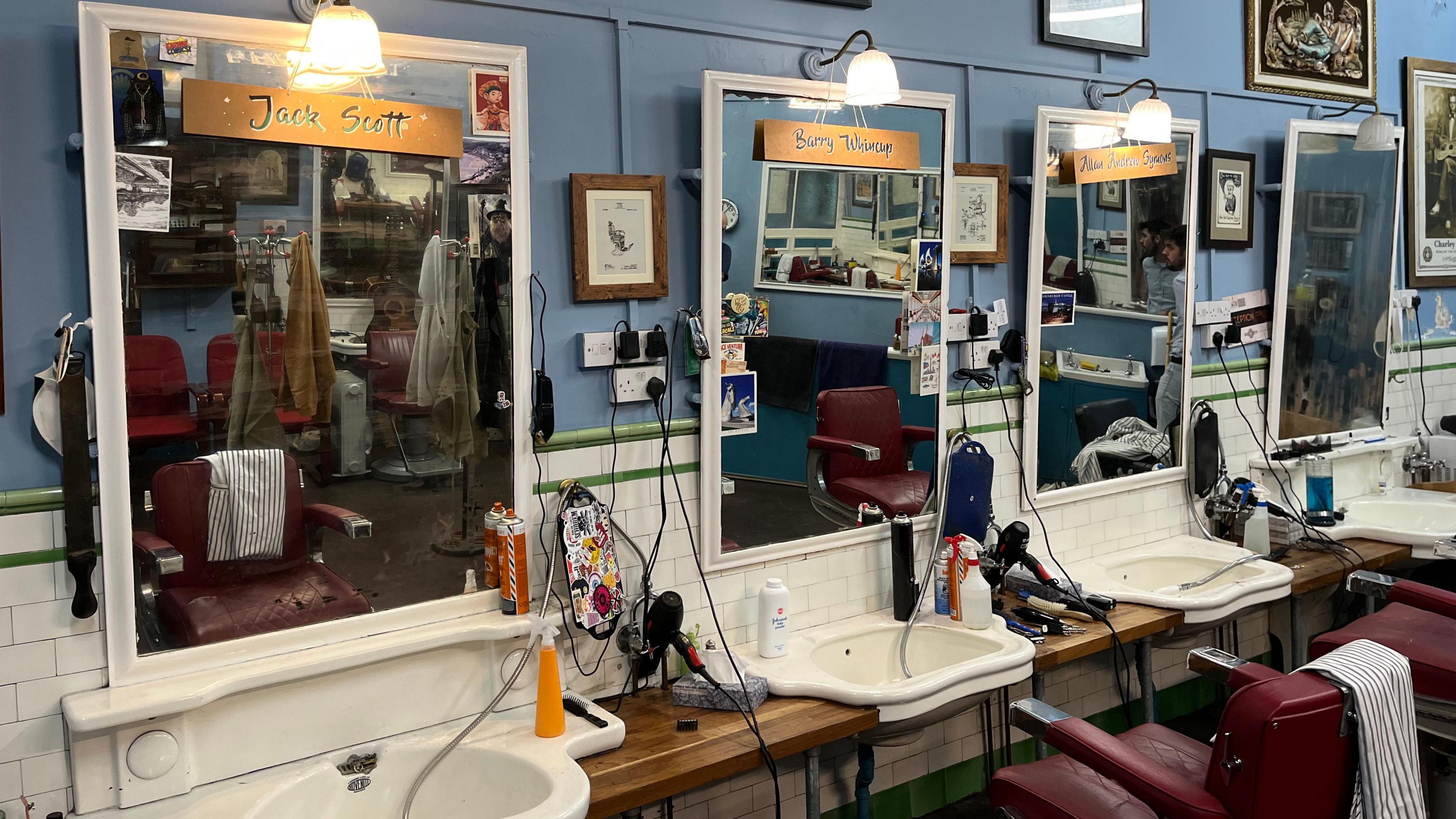 The inside of the barber shop. It shows four sinks and mirrors with traditional red barber chairs. There are hairdryers and hair cutting equipment. The names of each barber are on signs on the mirrors. A light is above each mirror. The wall has old-style white tiles around part of it.
