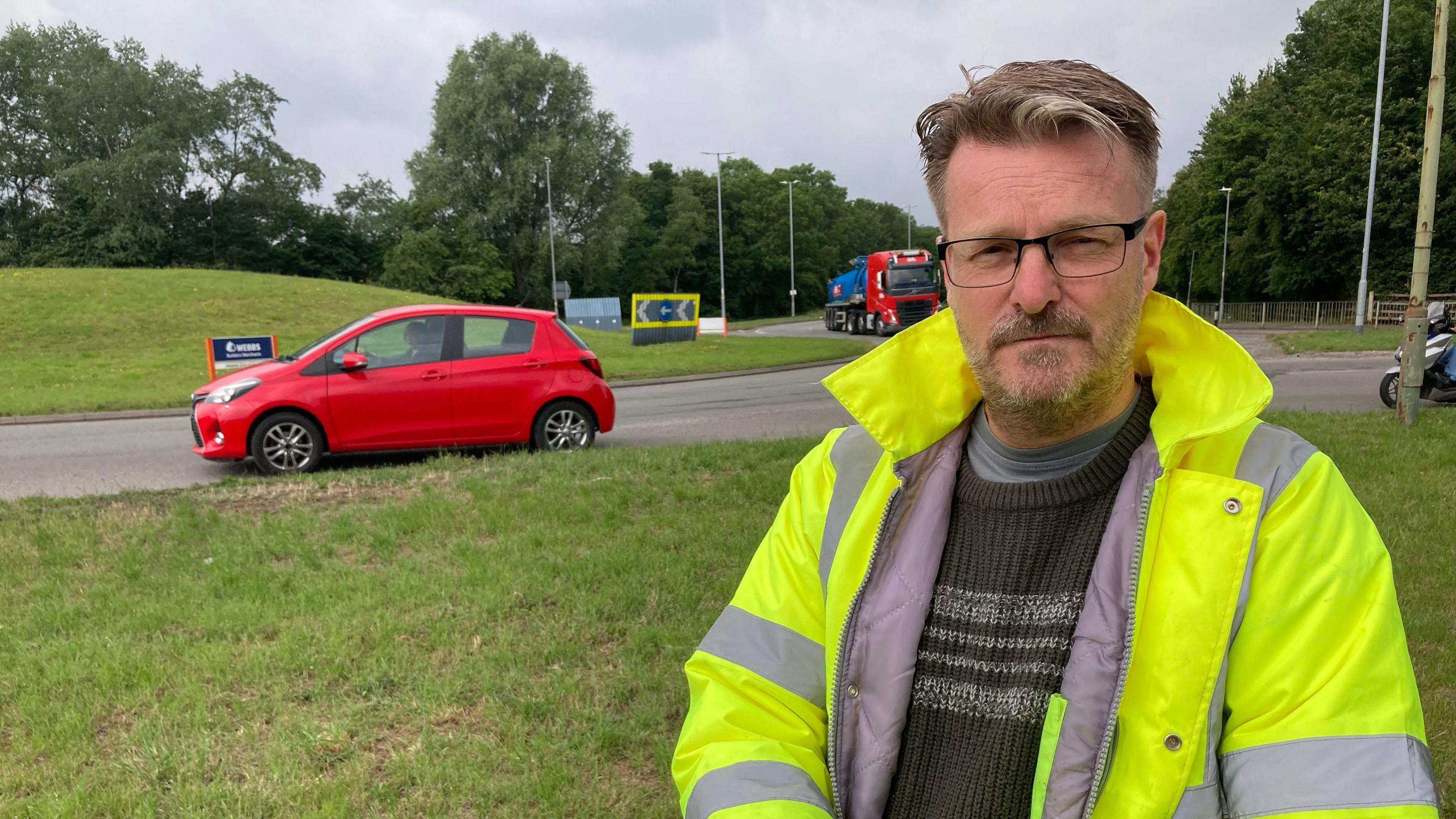 A middle aged man wearing glasses and a high-vis jacket stands in front of a roundabout with cars and road signs in the background.