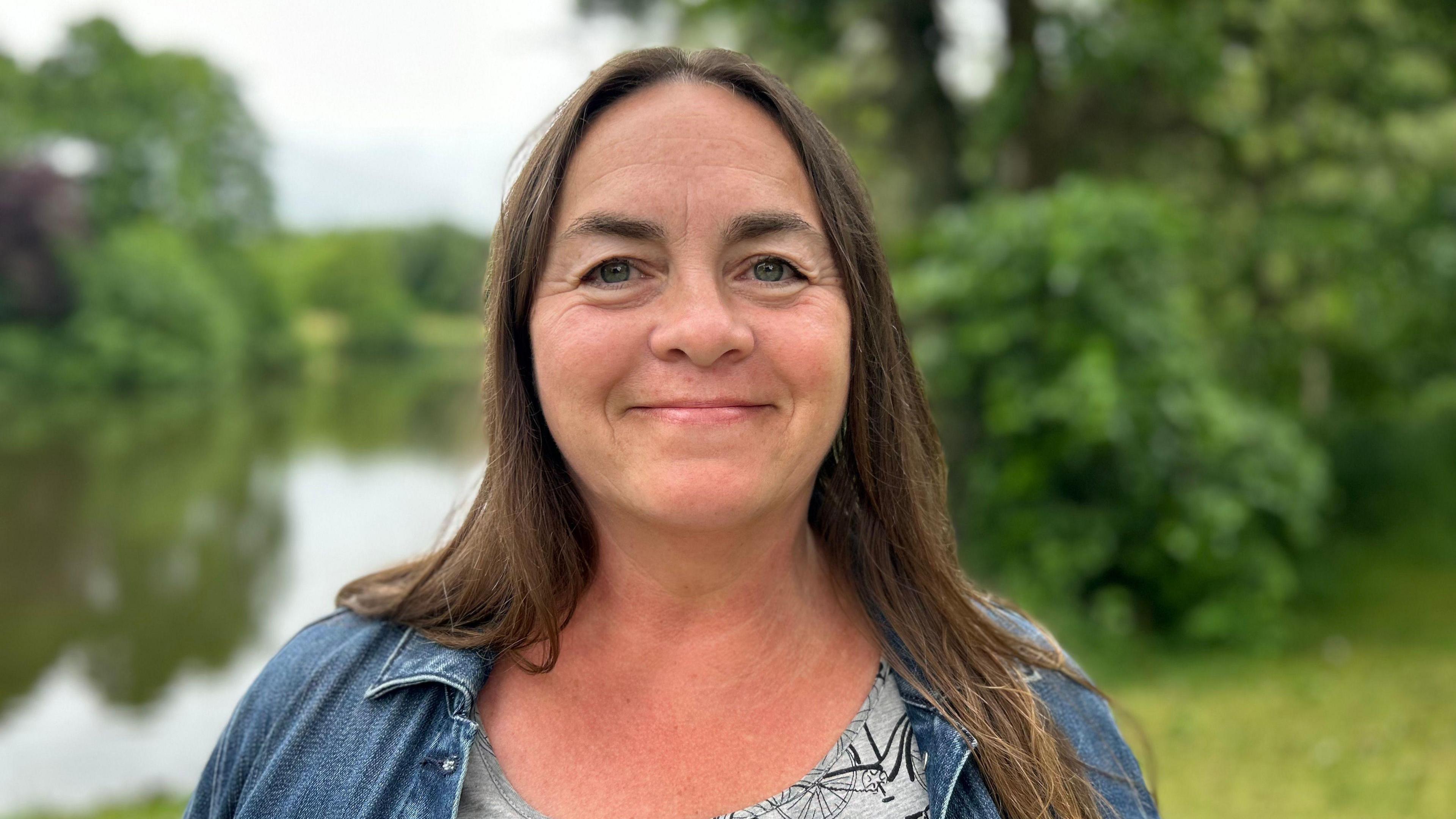 Diane, with brown hair and a blue denim jacket, standing in front of a river