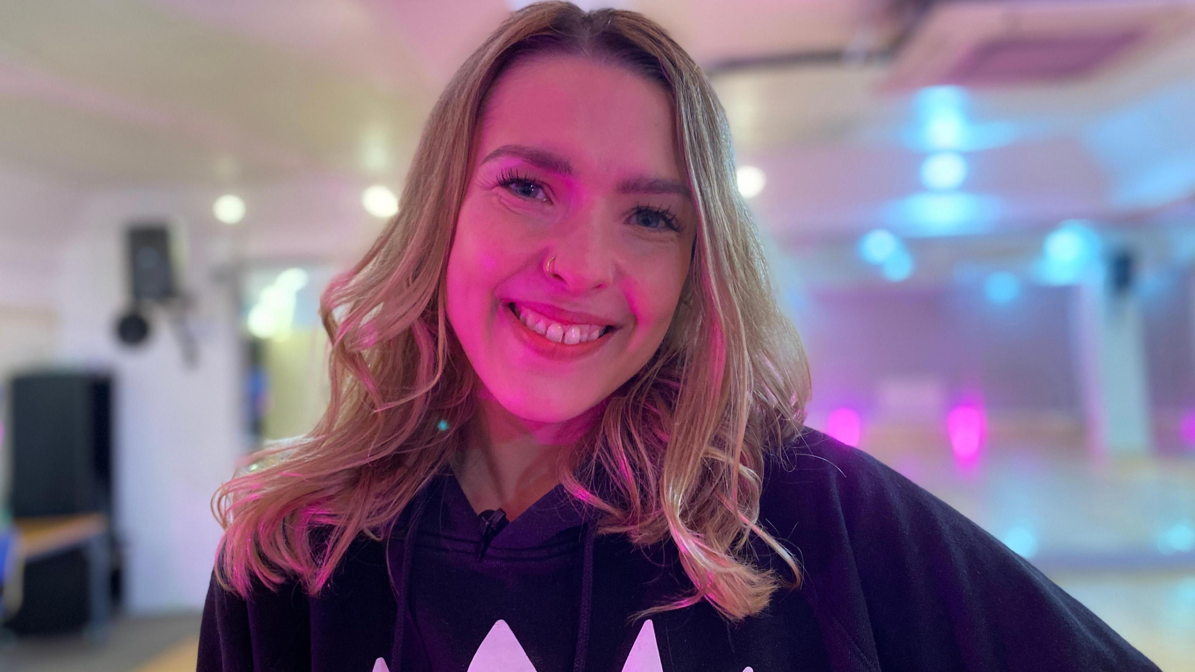 Amanda Radek has blonde shoulder-length hair and is wearing a black hoodie that says SOS. She is smiling at the camera. In the background there is a dance studio with pink and blue lights