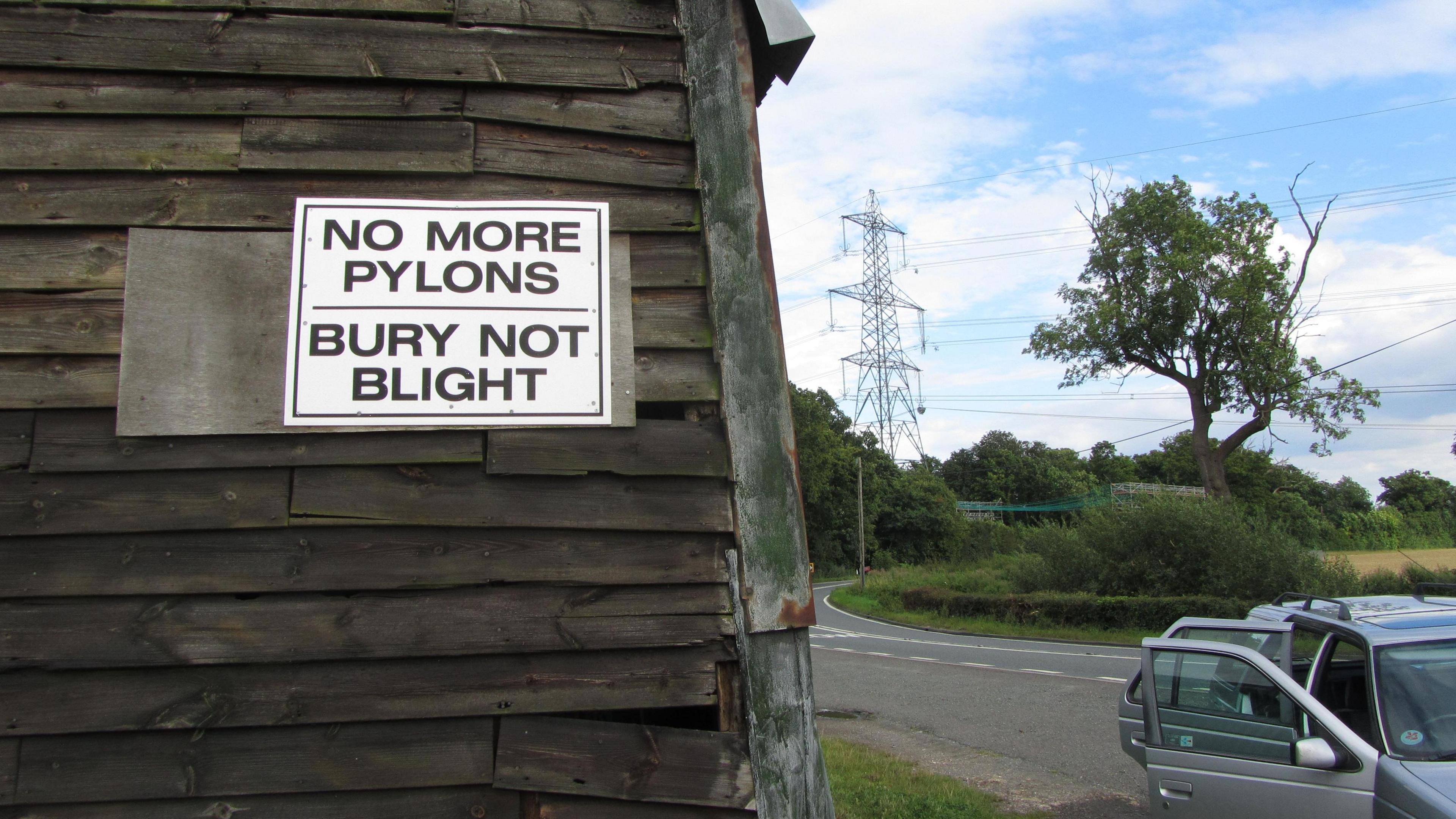 Sign on the side of a barn ("NO MORE PYLONS; BURY NOT BLIGHT") with a road and an existing electricity pylon in the background