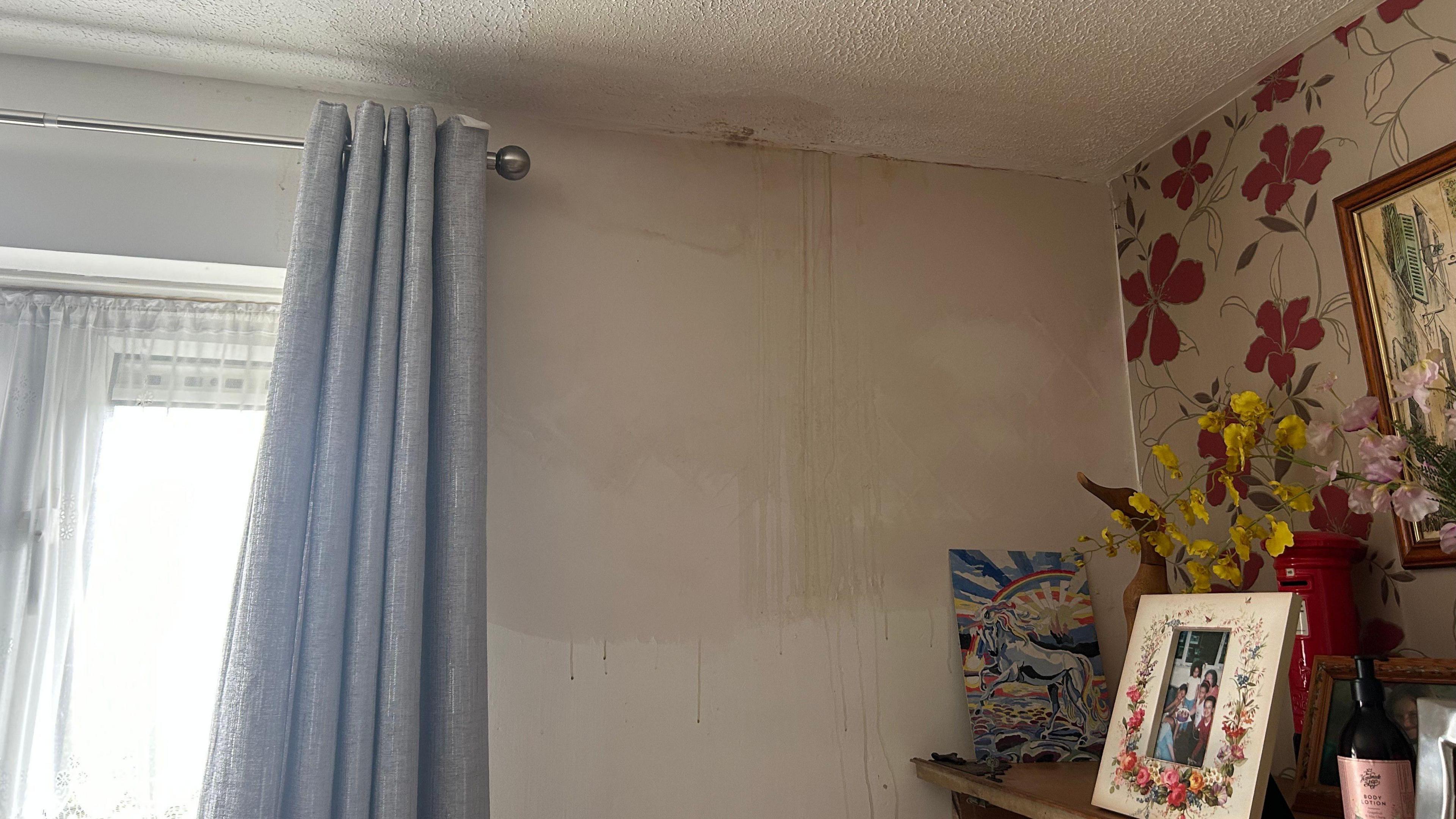 The inside of Brenda Gleeson's living room where damp drips through the ceiling on to the wall. On the left there is red flower wallpaper and a mantlepiece with fake flowers and picture frames.