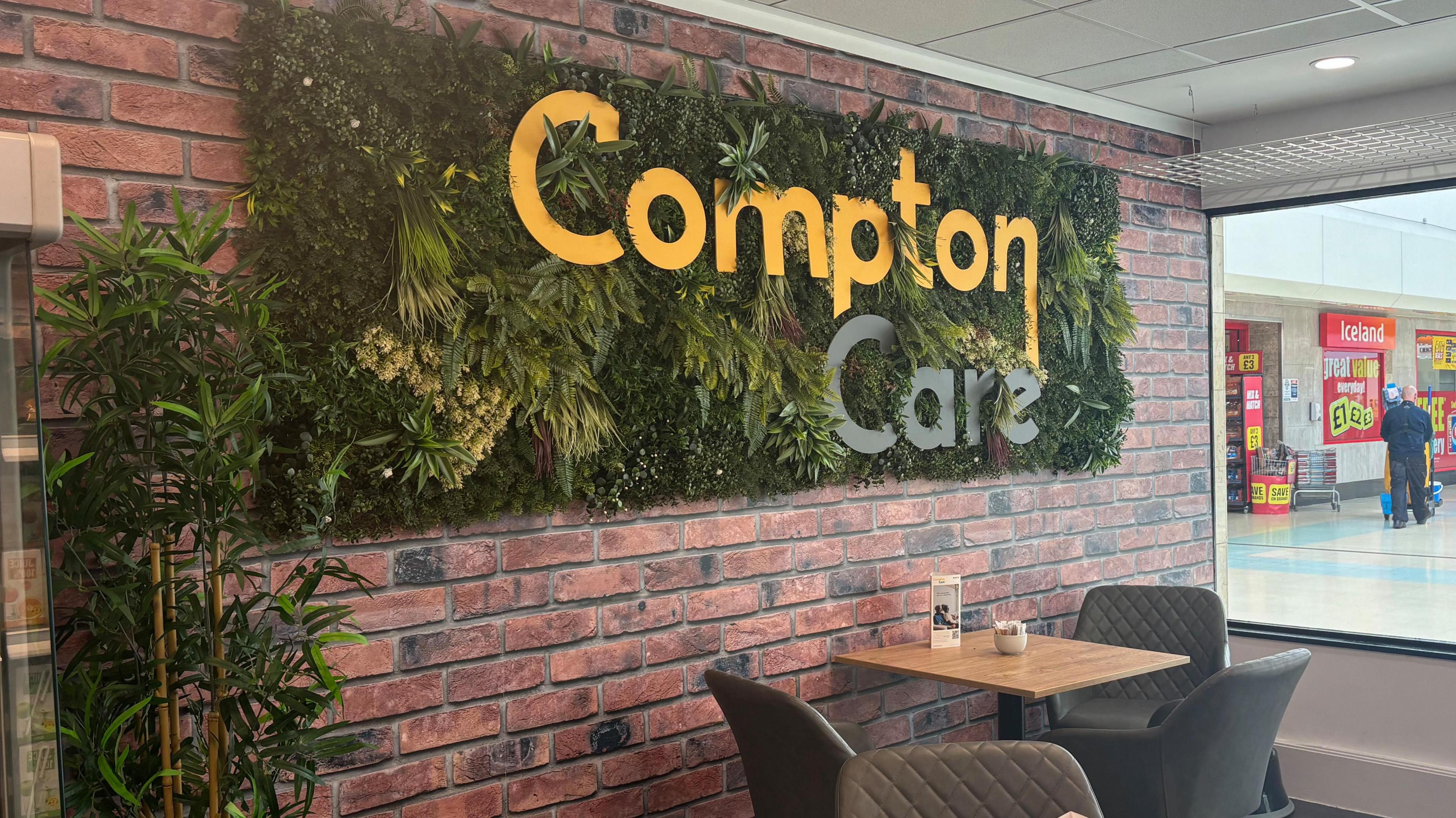 Compton Care cafe sign, on a wall of plants. The sign is yellow and Grey. Grey cafe seats are also in the image. 