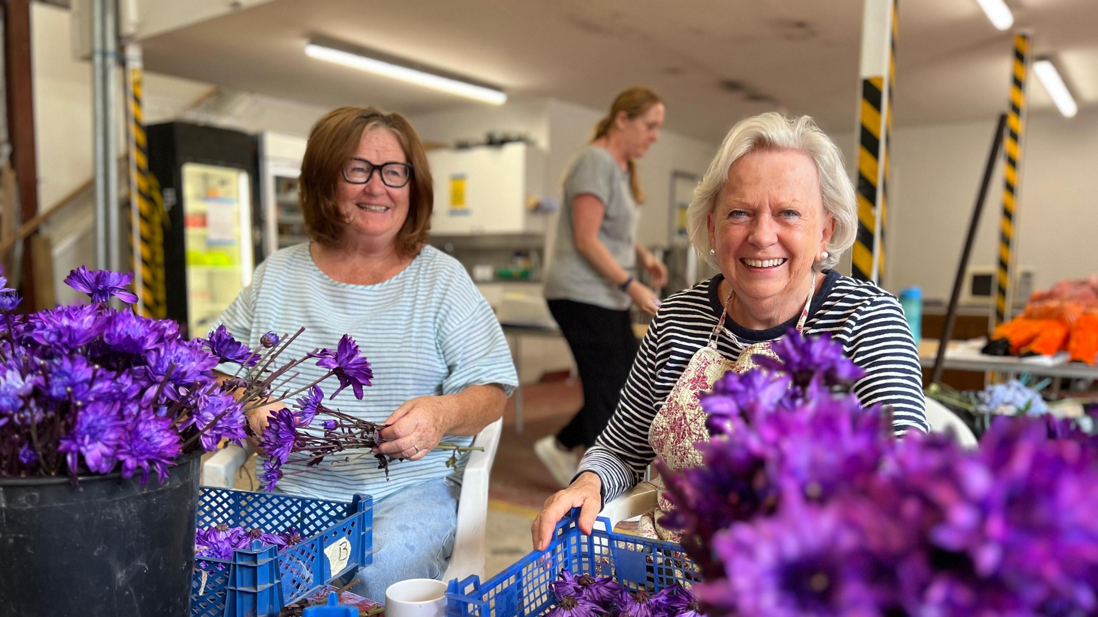 Two women smiling, sat down with crates of purple flowers on their laps and purple flowers in front of them in the foreground. Another woman is out of focus behind them walking past