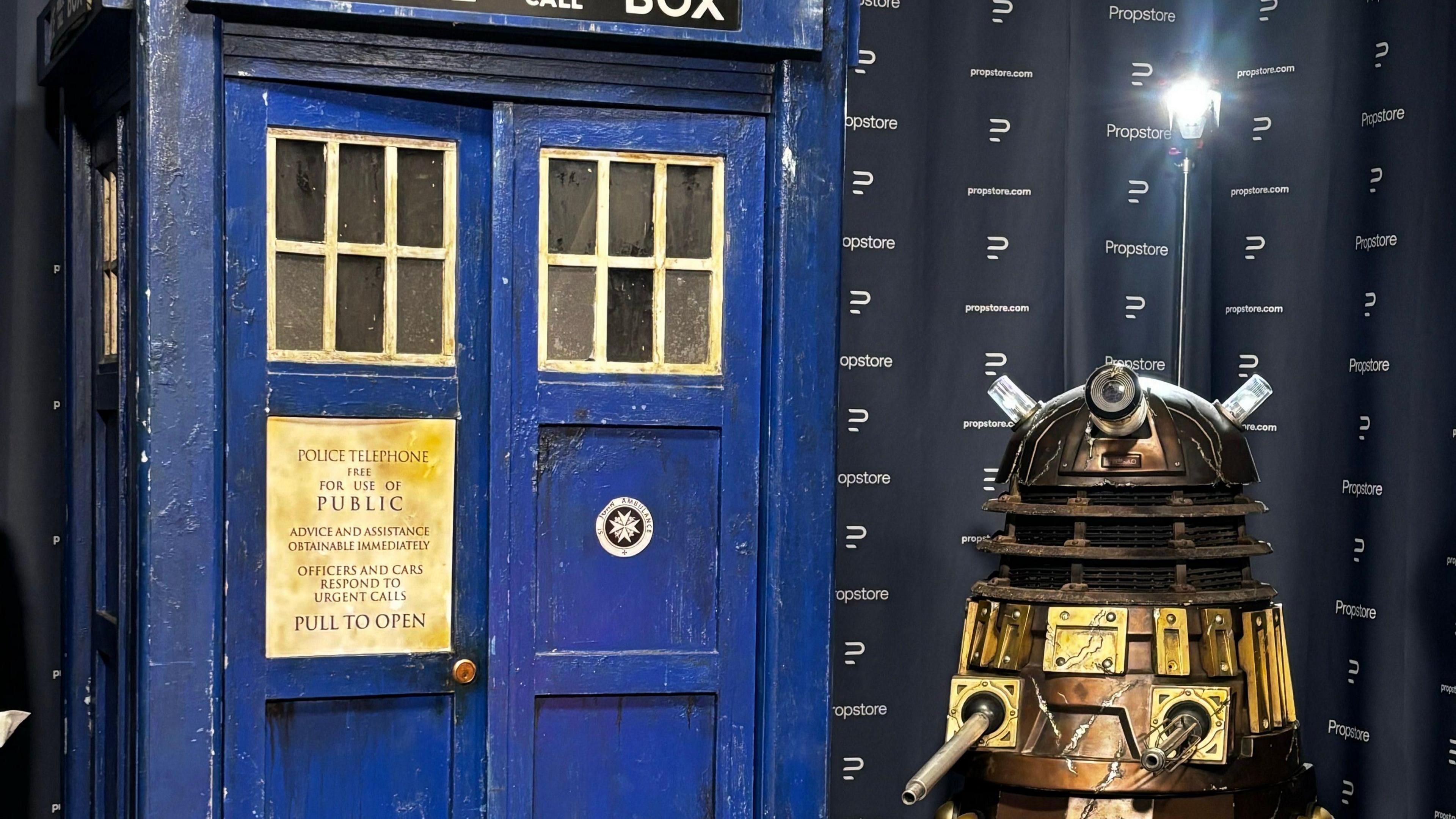 A blue TARDIS is sat next to a bronze and scarred Dalek