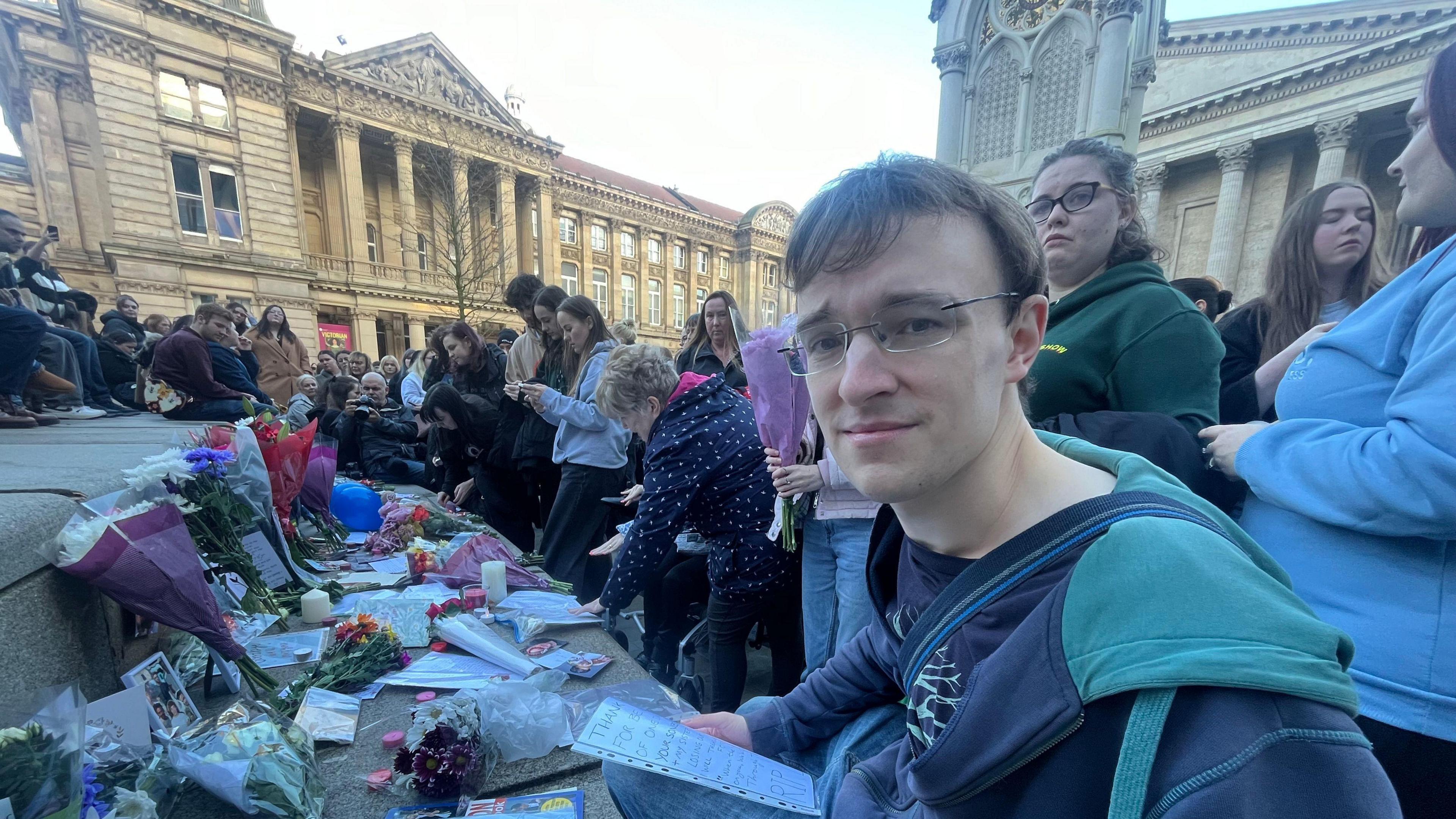 A young man kneels in front of a set of steps which are covered in bouquets of flowers and written tributes. He wears a blue jacket and black glasses, while holding a piece of paper in his hand and looking solemnly at the camera. Crowds of people are around and behind him. A large building with pillars is behind them.