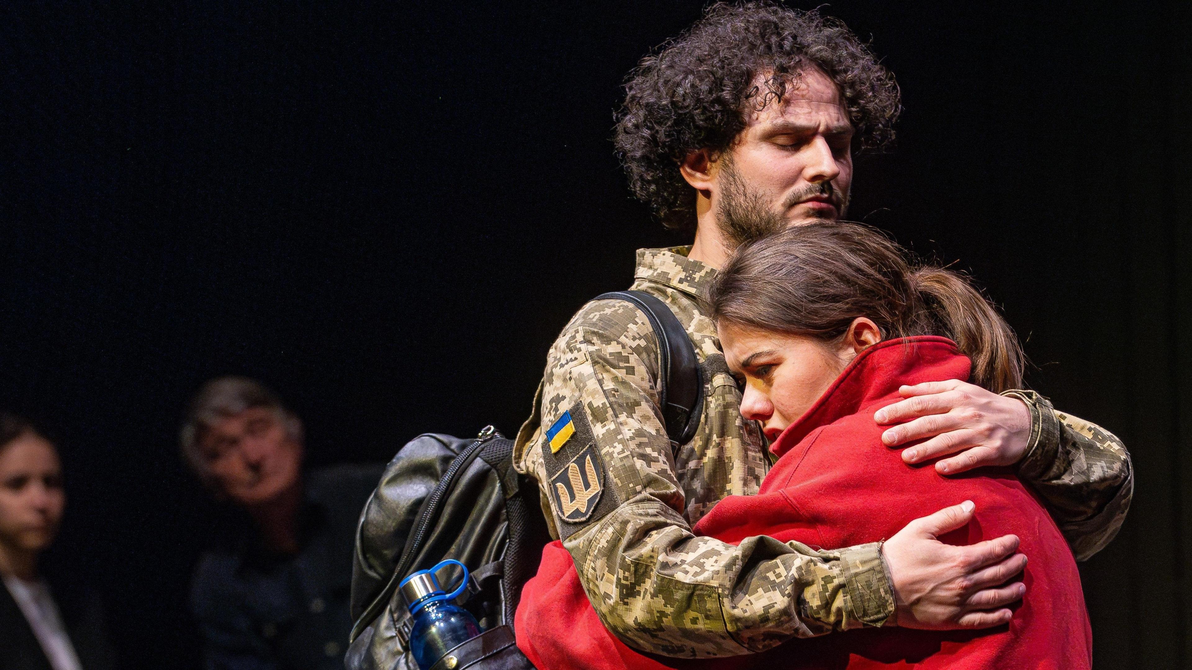 A male actor with curly hair and a beard, dressed in military uniform, embraces a brown-haired female actor wearing a red jumper. In the background, two audience members can be seen watching.