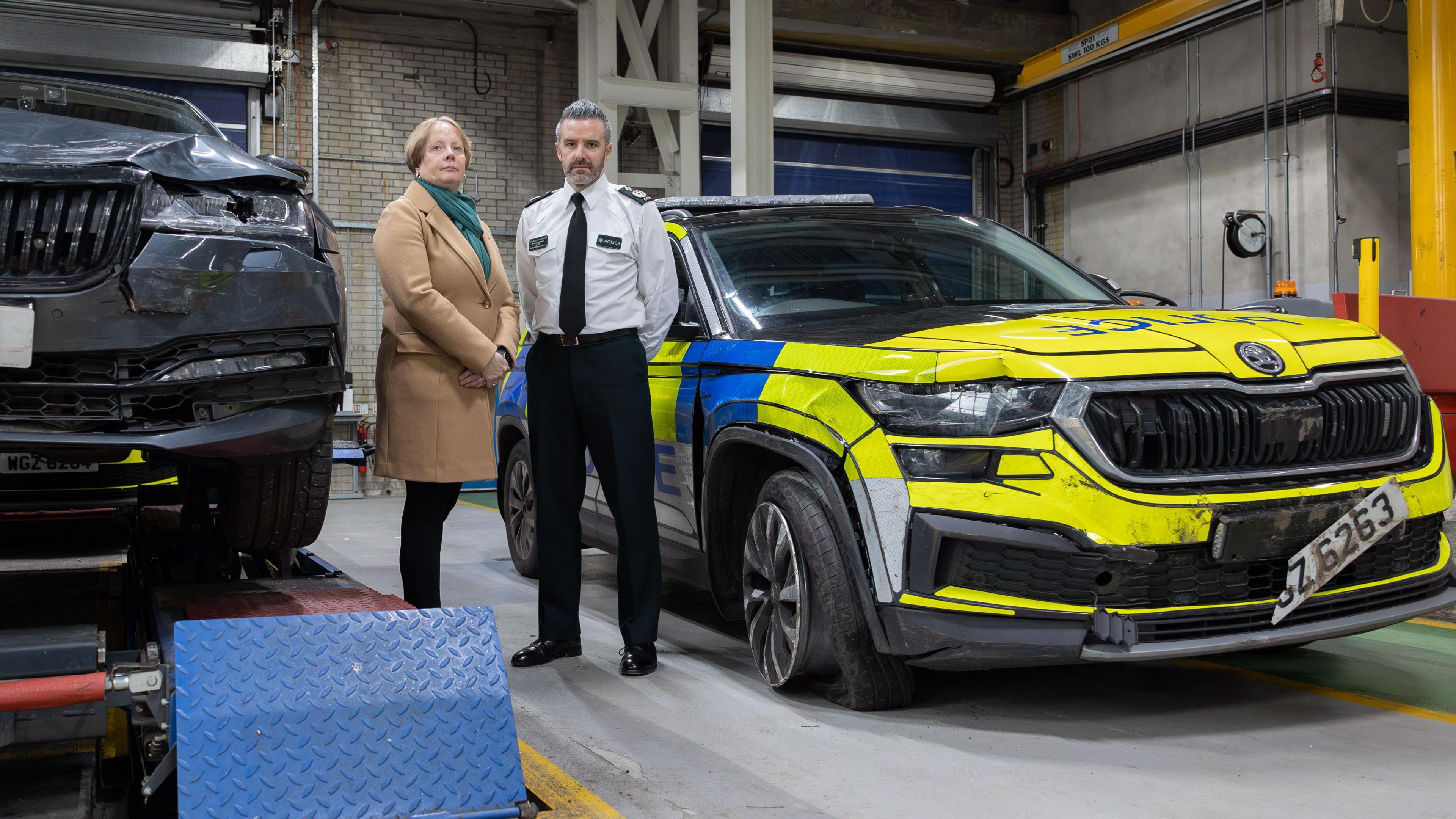 A woman is wearing a brown coat. An officer is next to her. There are damaged police cars beside them.