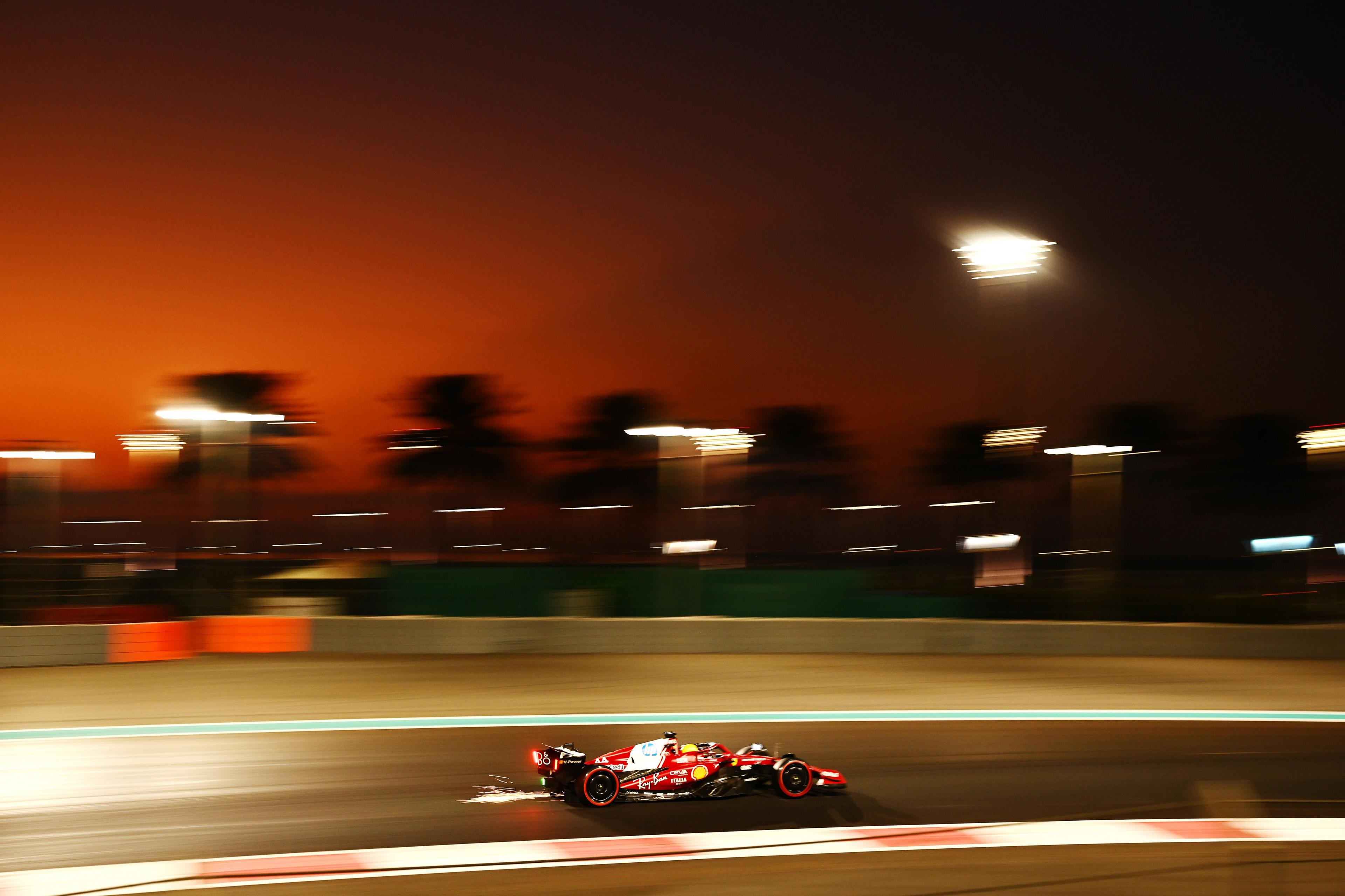 Un coche rojo de Fórmula 1 avanza a toda velocidad por la pista al anochecer, con destellos que salen de la parte trasera. Palmeras borrosas y focos brillantes se recortan contra un cielo naranja y morado intenso.