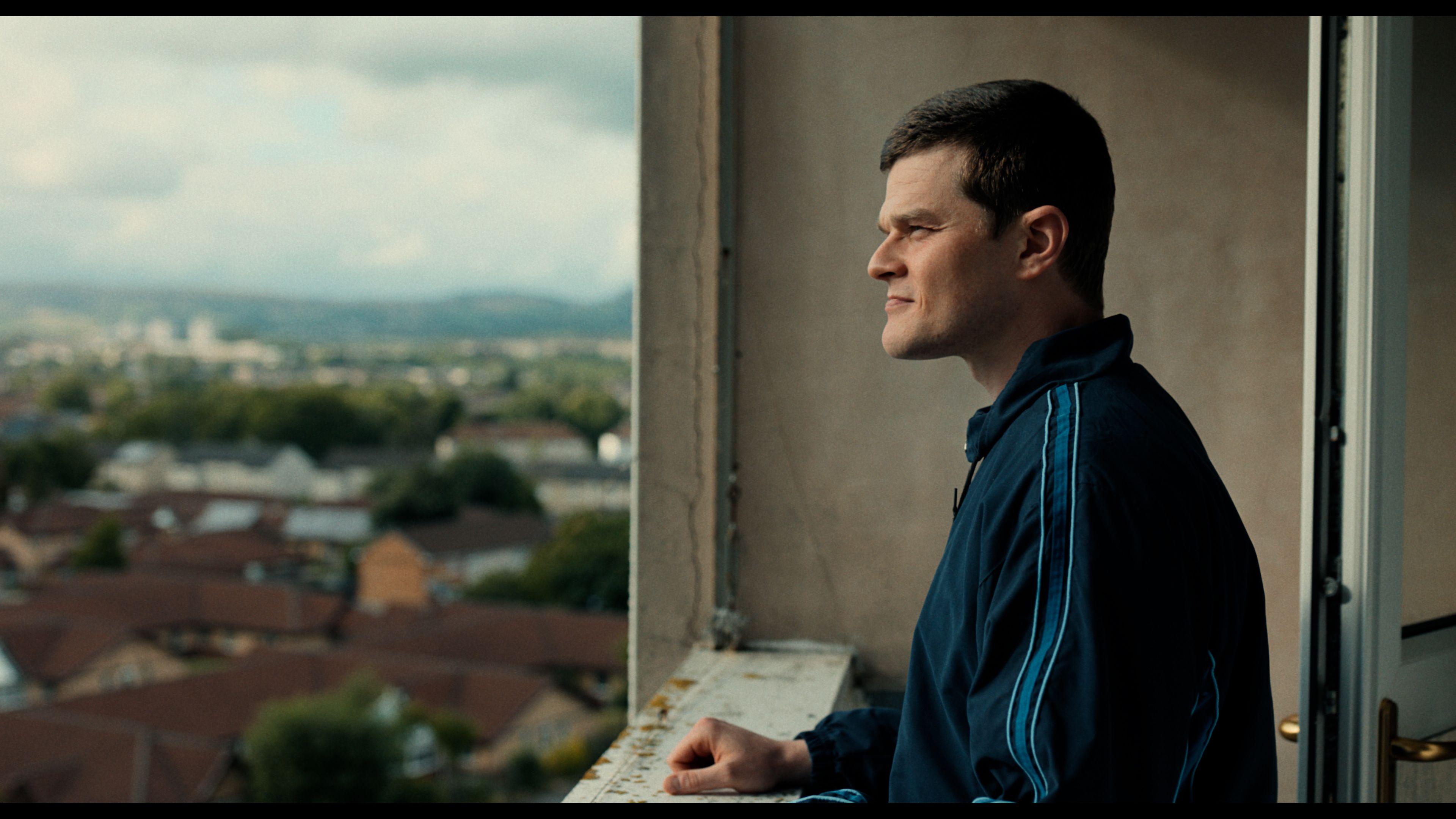A photo from the film of the actor Robert Aramayo on the balcony of a high flat looking out. He has a close cropped hair cut and is wearing a navy and blue sweatshirt. 