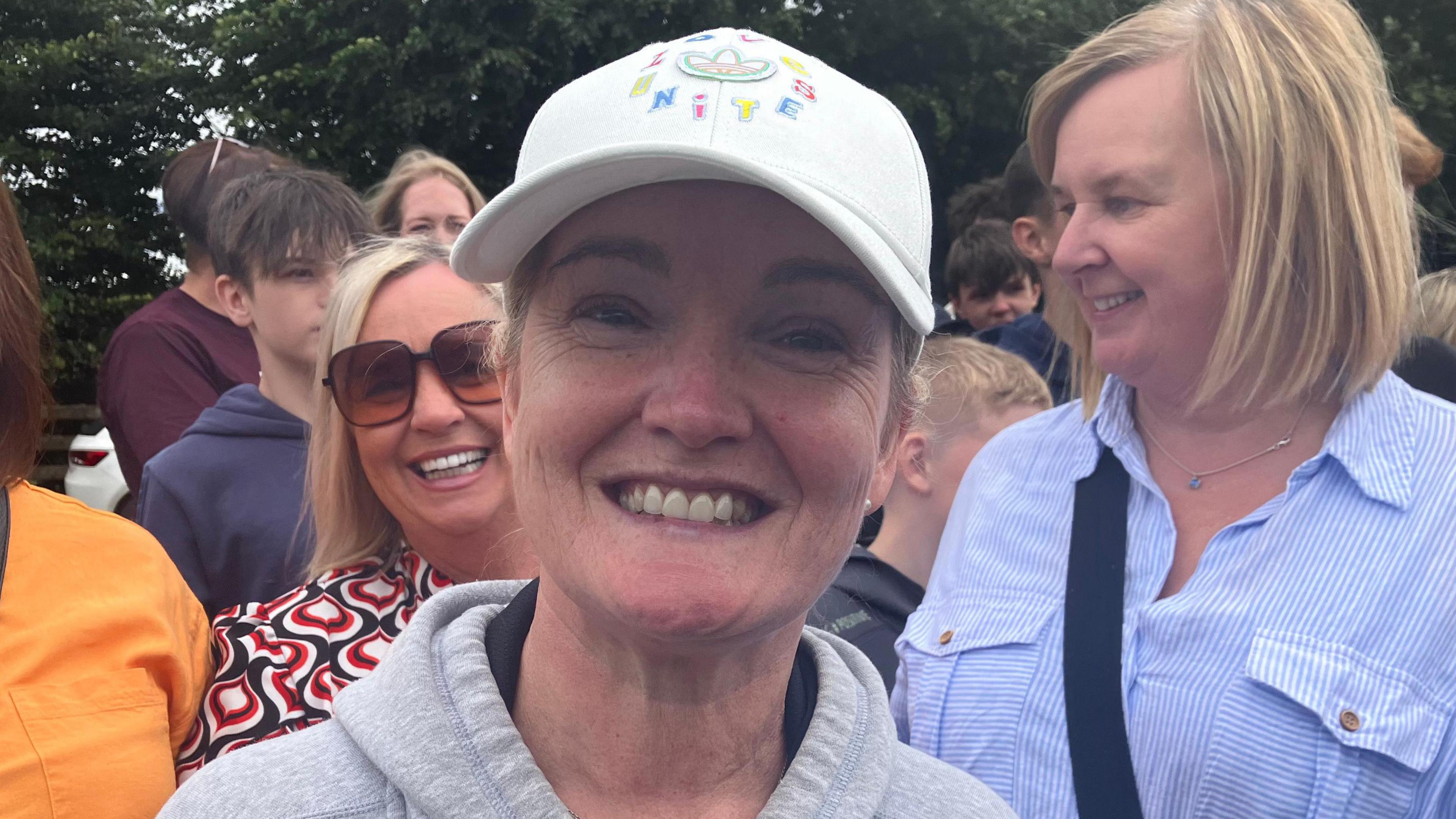 Alicia O'Kane smiling broadly, while wearing a grey hoodie and a white baseball cap. There are other onlookers in the background smiling and having a good time..